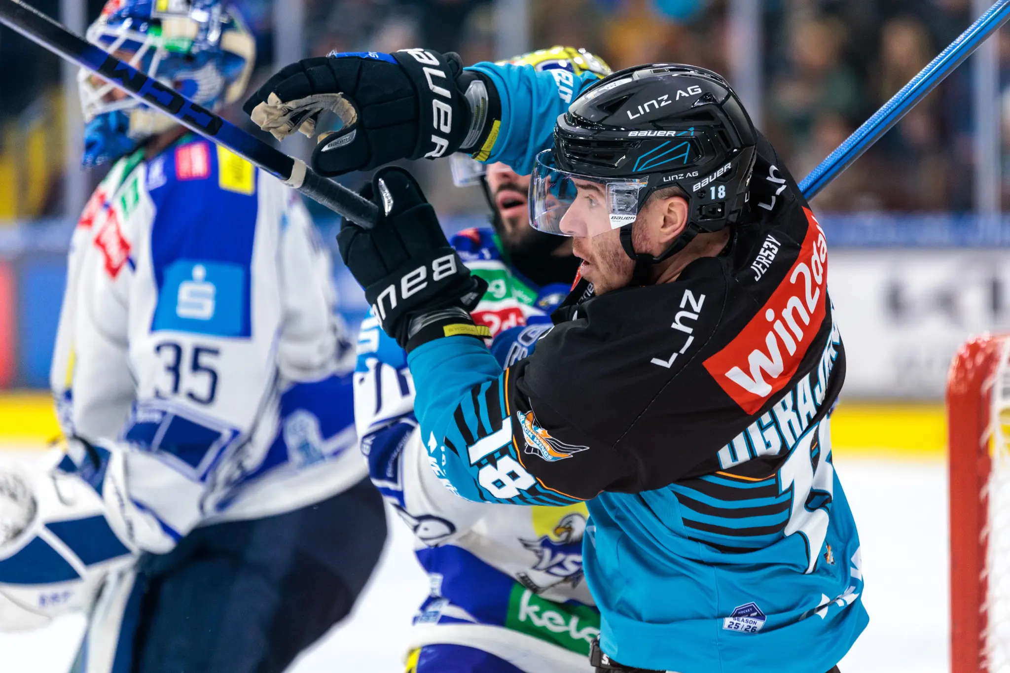 Ein Eishockeyspieler der Black Wings Linz im schwarzen Trikot und ein VSV-Torhüter im blau-weißen Trikot sind in einem intensiven Stockschlag-Duell auf dem Eis