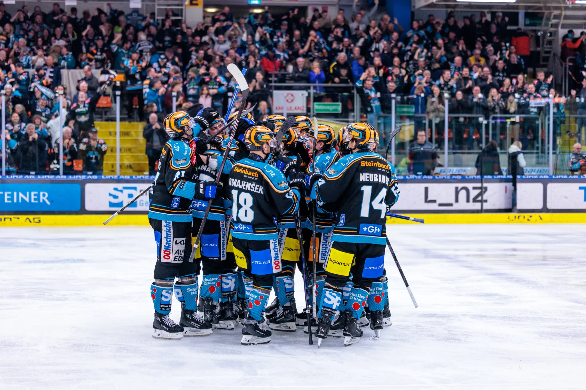 Eine Gruppe Eishockeyspieler der Black Wings Linz in ihren schwarz-hellblauen Trikots jubeln eng beisammen auf dem Eis 