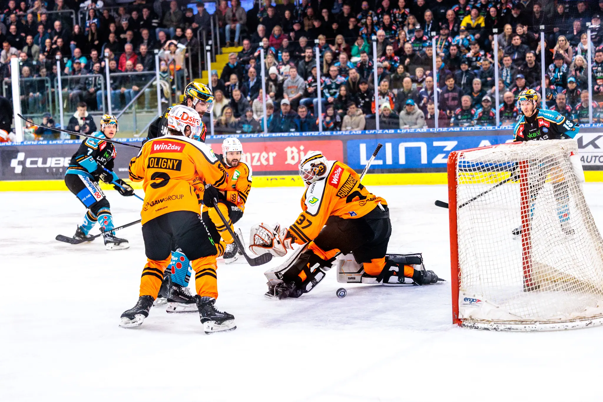 Eishockey-Torwart in orangefarbener Ausrüstung kniet in Spielposition vor dem Tor, umgeben von Feldspielern, während der Puck durch seine Beine ins Tor wandert 