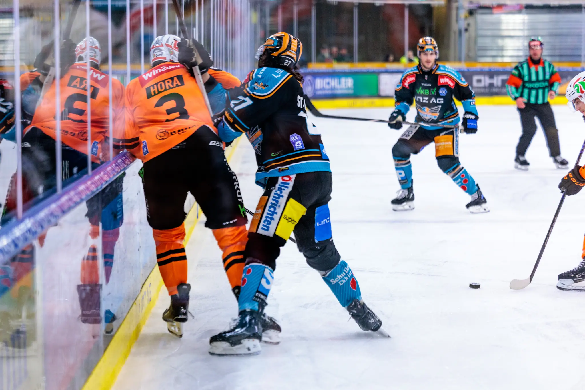 Zwei Eishockeyspieler in orangefarbenen und dunkelblauen Trikots kämpfen an der Bande um den Puck 