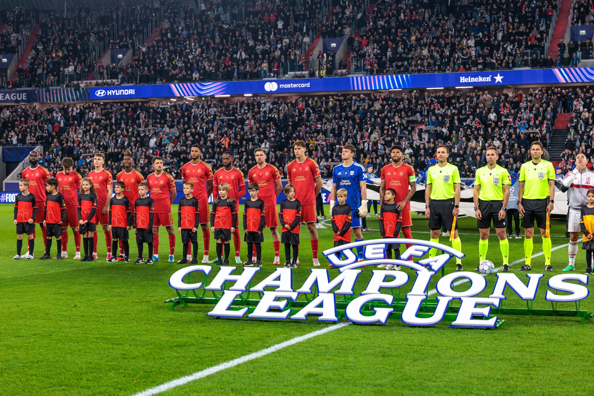 Die LASK-Mannschaft steht in roten Trikots mit Begleitkindern und dem neongelben Schiedsrichtertrio vor dem Champions-League-Logo auf dem Rasen.