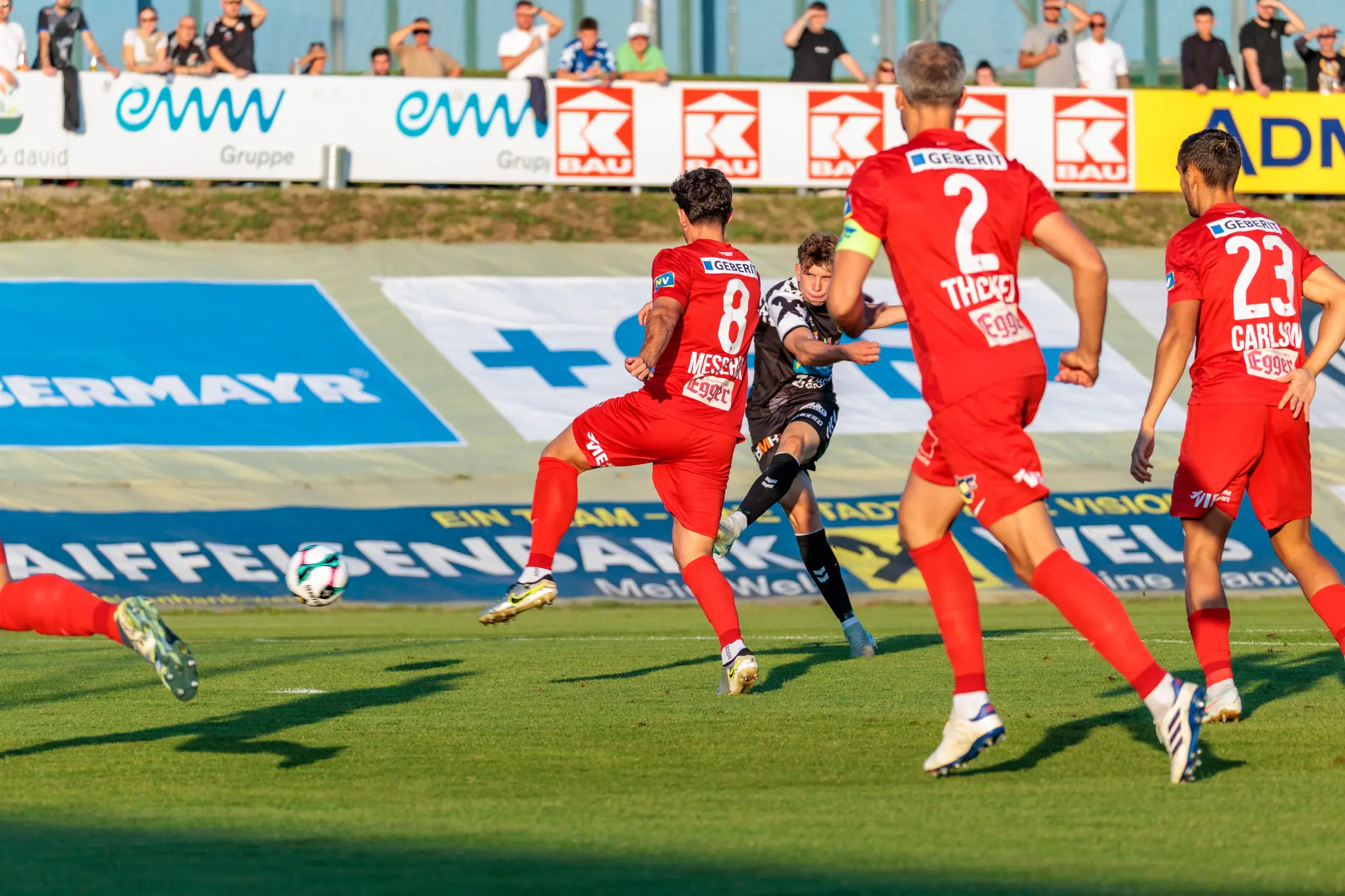 Ein Spieler der SPG HOGO Wels versucht im schwarz-weißen Trikot einen Schuss, der von mehreren Spielern des SKN St. Pölten in roten Trikots verdeckt wird