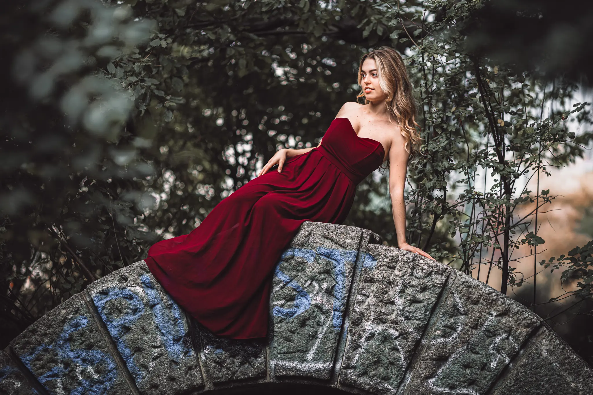 Model Valentina posiert elegant in einem langen, roten Kleid auf dem Bogen einer verwitterten Steinbrücke im Wiener Stadtpark
