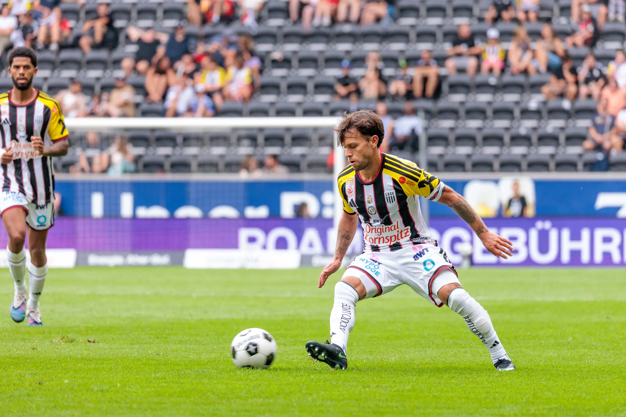 LASK-Spieler bereitet den Spielaufbau vor, das Düsseldorfer Tor ist im Hintergrund sichtbar, ein Mitspieler beobachtet die Szene.