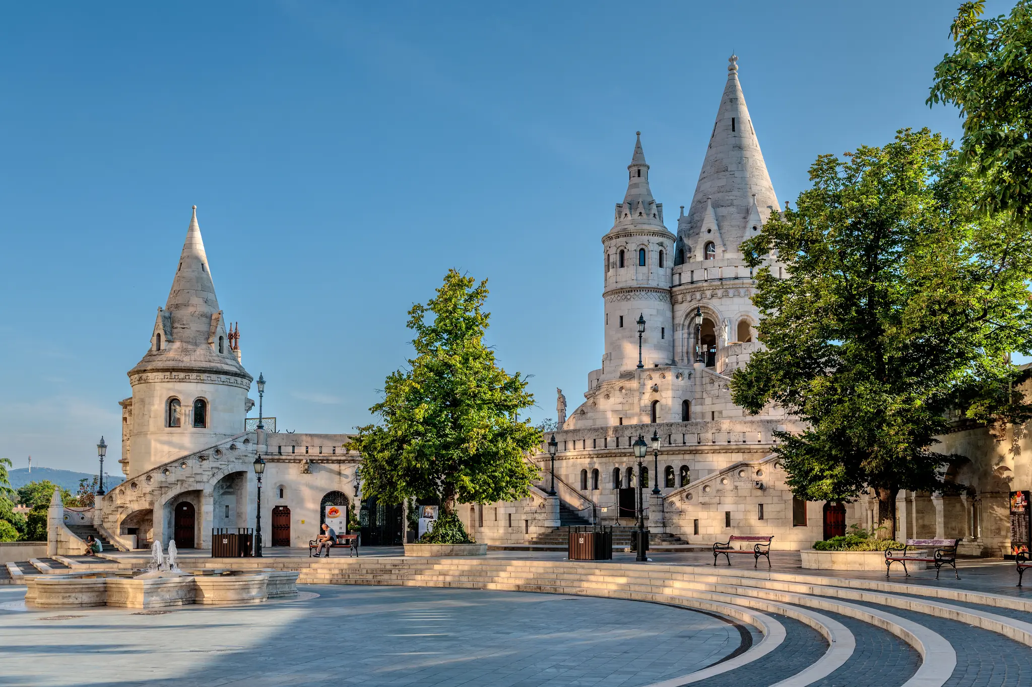 Die Fischerbastei in Budapest mit ihren markanten weißen Türmen und steinernen Treppen an einem sonnigen Tag. Im Vordergrund befindet sich ein gepflasterter Platz mit einem Brunnen und grünen Bäumen unter blauem Himmel. 