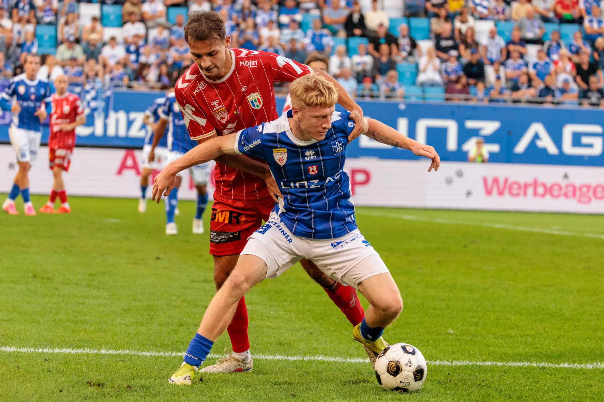 Ein Welser Spieler greift von hinten nach dem Linzer Angreifer, der den Ball am Fuß führt; im Hintergrund leuchtet die Linz-AG-Bande.