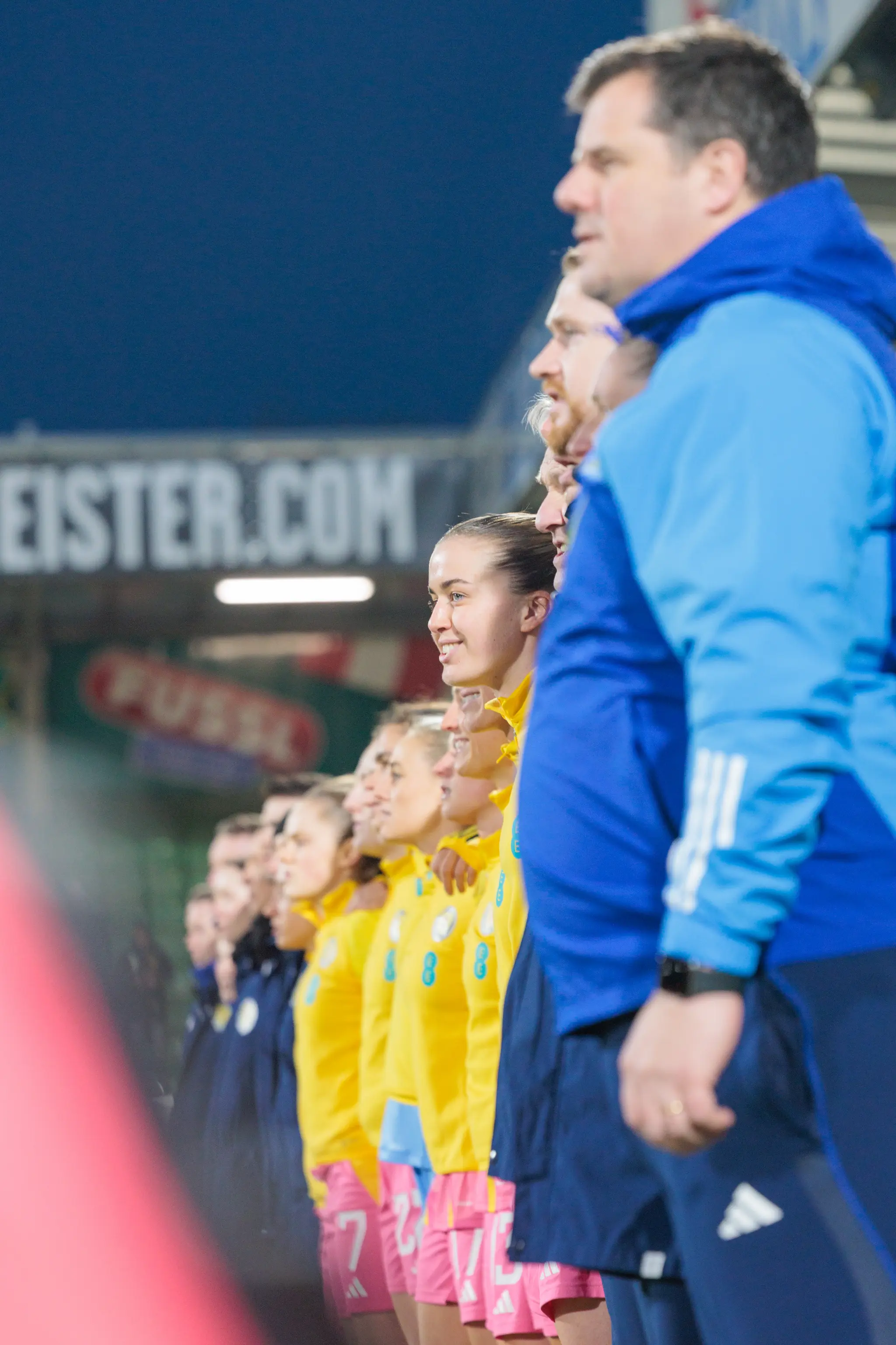Eine junge Fußballspielerin in gelbem Trikot und pinker Hose lächelt, umgeben von ihrem Team und Trainern, die in einer Reihe in einem Stadion stehen