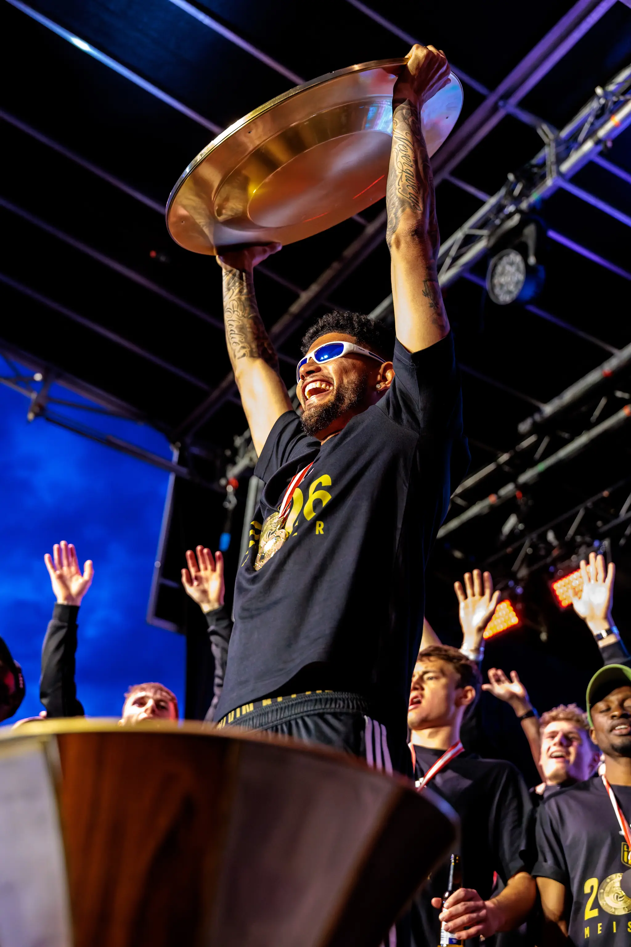 Ein Spieler mit Sonnenbrille jubelt auf der Bühne und hält den goldenen Meisterteller hoch