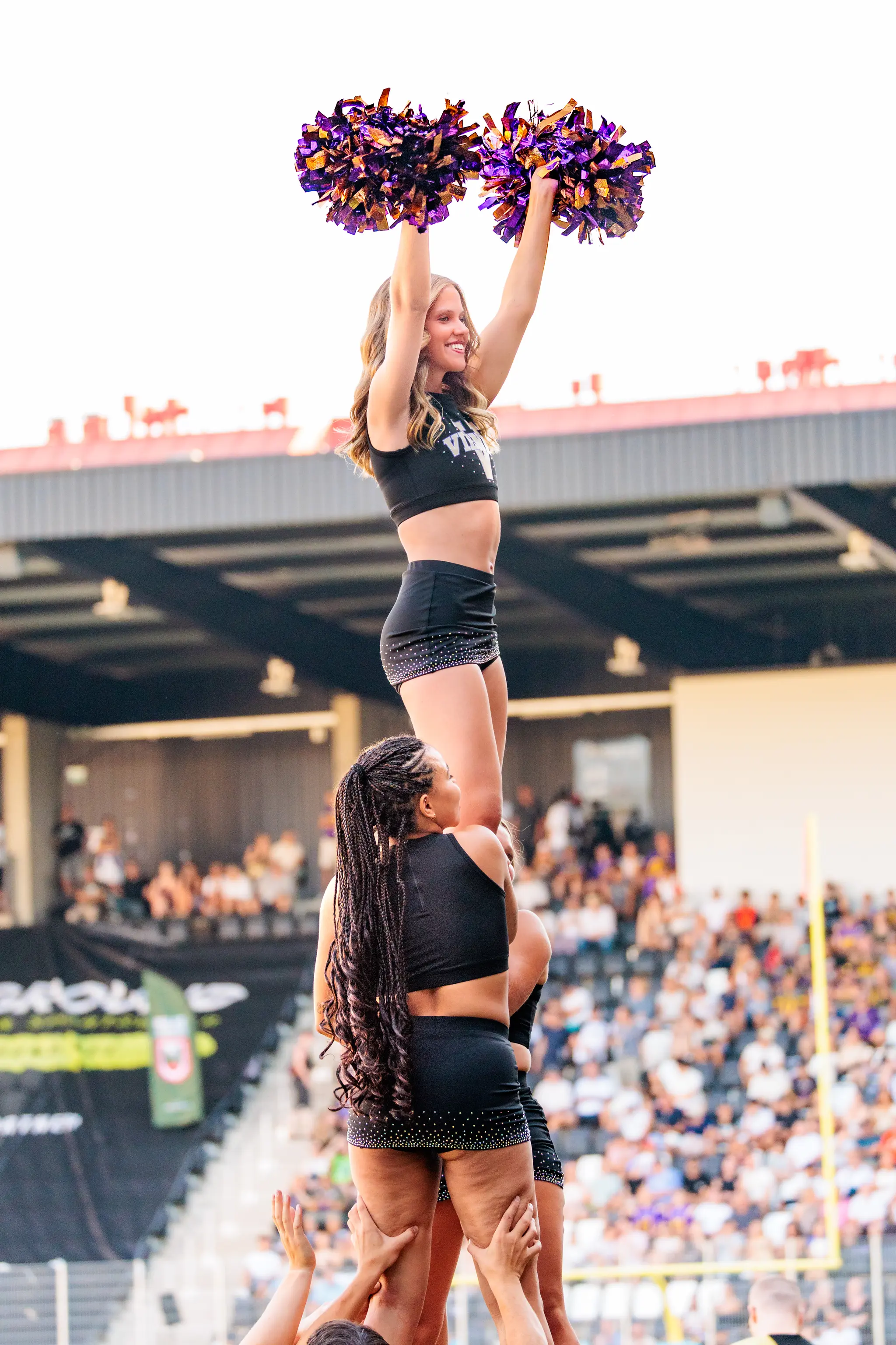 Cheerleaderin der Vienna Vikings wird bei einer Stunt-Figur hochgehoben und schwingt violett-goldene Pompons.