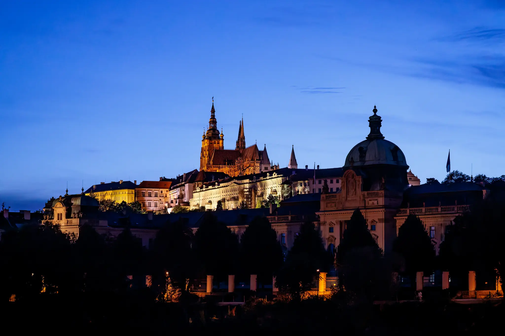 Die beleuchtete Prager Burg mit den Türmen des Veitsdoms ragt über der dunklen Baumreihe der Kleinseite auf; in der blauen Stunde tragen die Stadtlichter die Abendstimmung über die Moldau.