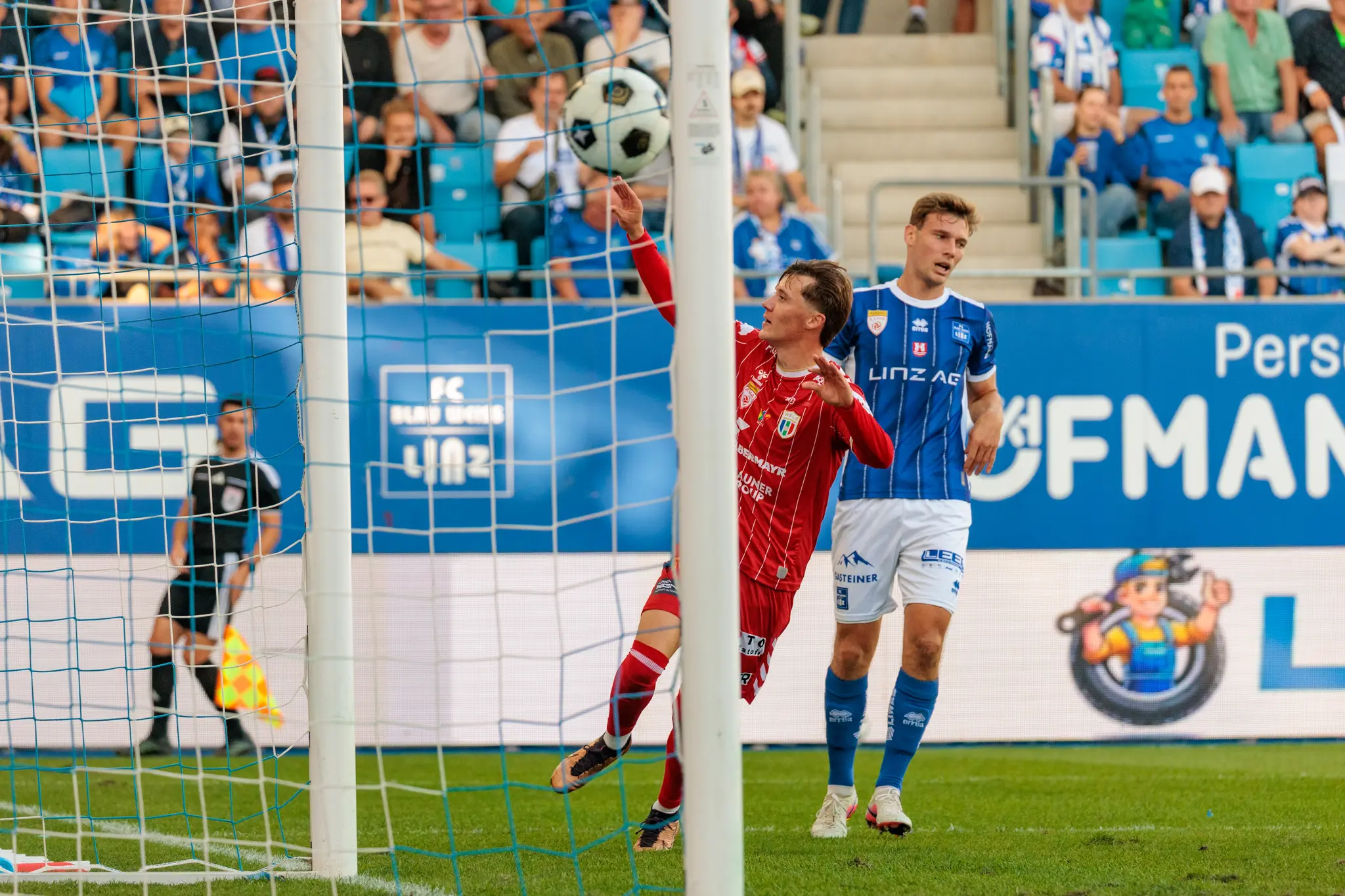 Der Ball hängt im Tornetz – der Führungstreffer der Welser ist gefallen; Torschütze Sebastian Malinowski richtet daneben eine freche Handgeste an die Heimtribüne, ein Linzer Spieler blickt zu.