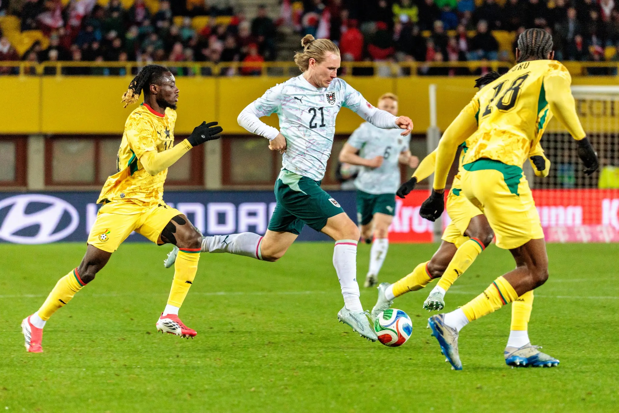Ein österreichischer Fußballspieler im weißen Trikot dribbelt den Ball während eines Länderspiels gegen Ghana, flankiert von zwei ghanaischen Gegenspielern in gelben Trikots