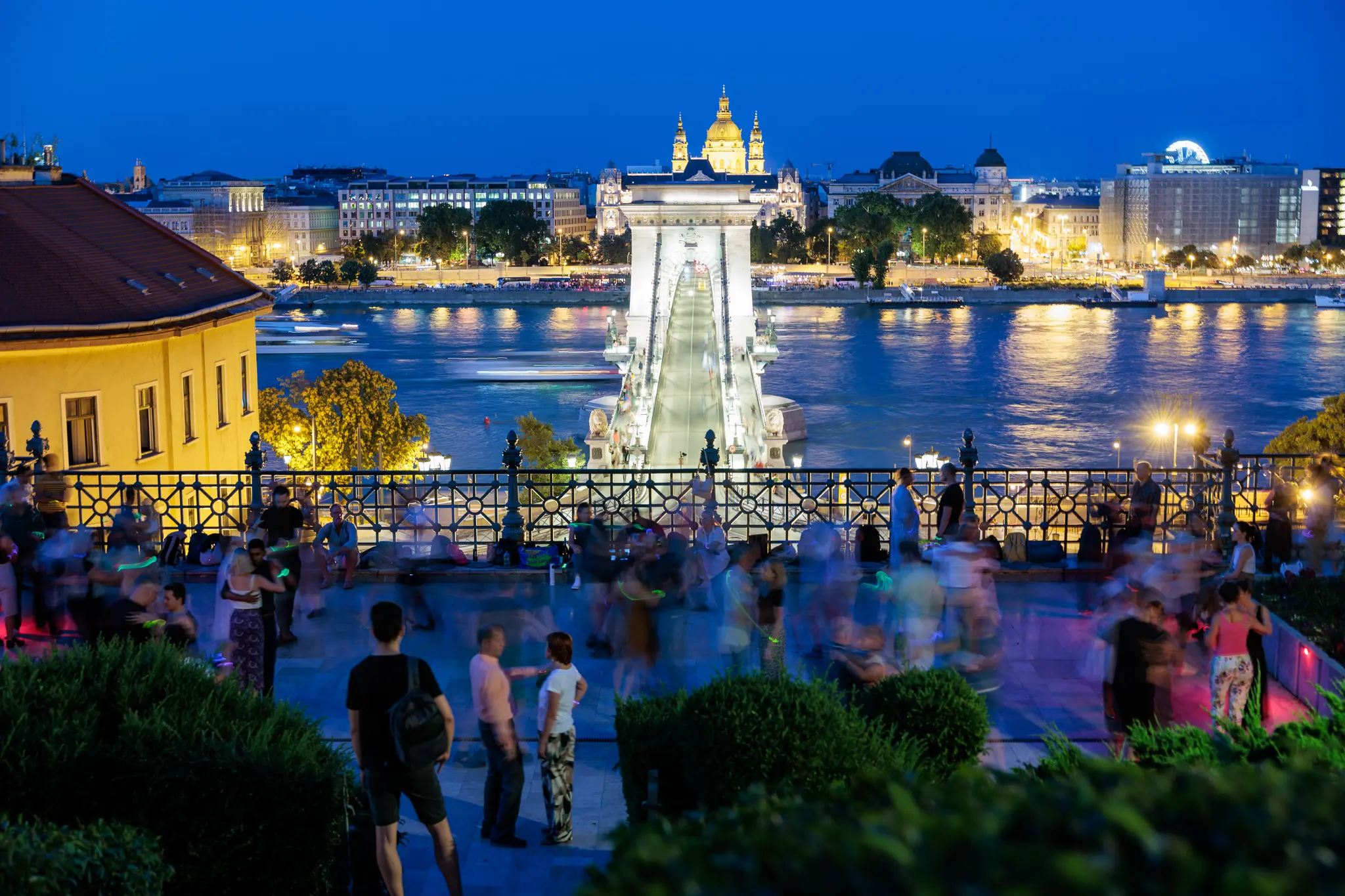 Nachtaufnahme der beleuchteten Széchenyi-Kettenbrücke in Budapest über die Donau, mit einer tanzenden Menschenmenge bei einem Outdoor-Event am Flussufer und der beleuchteten Stadtsilhouette im Hintergrund. 