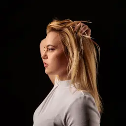Junge blonde Frau im Profil mit hellem Oberteil, die ihr Haar vor dunklem Hintergrund berührt