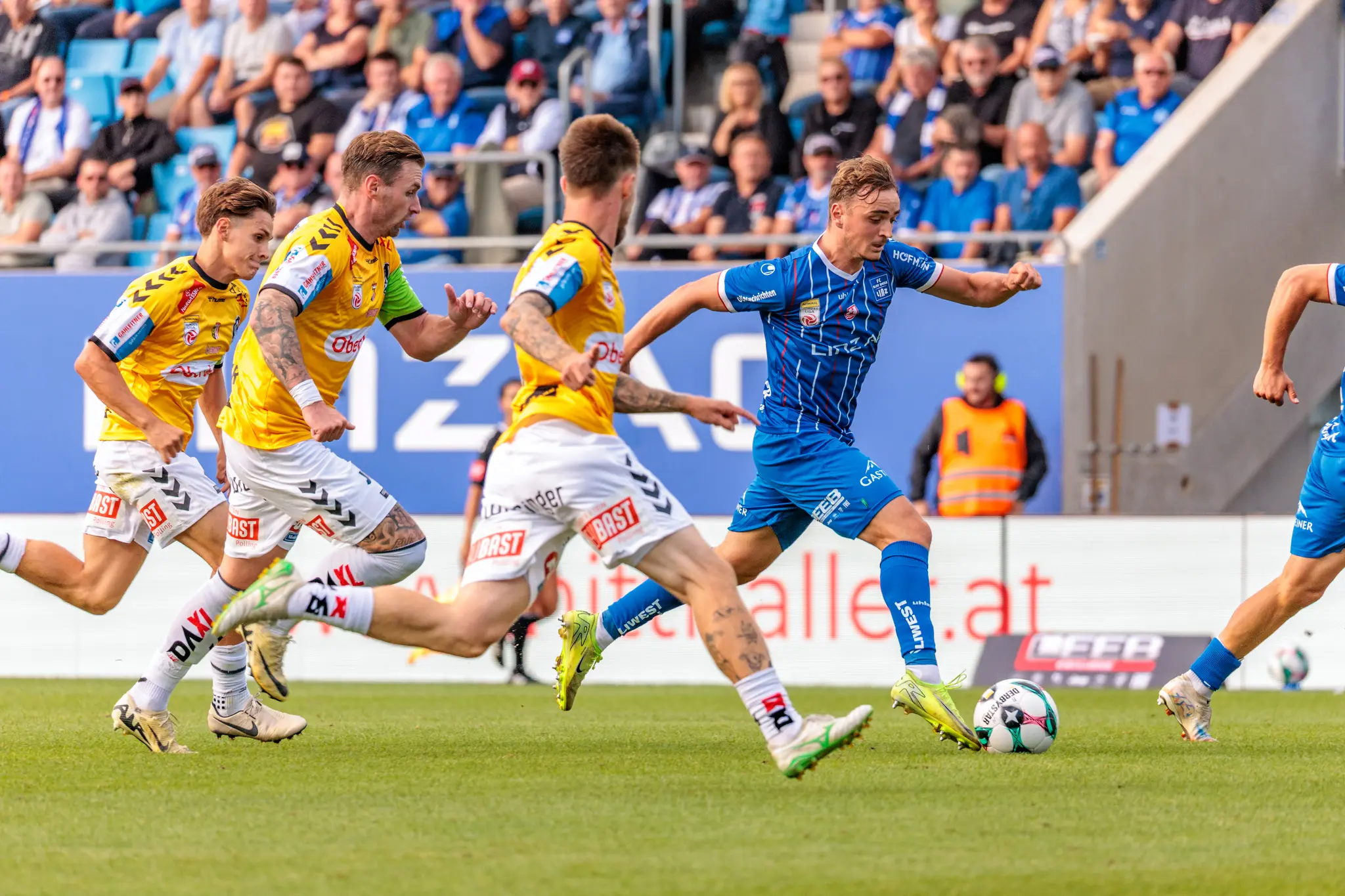 Ein Spieler des FC Blau-Weiß Linz im blauen Trikot versucht einen Torschuss auf dem Fußballfeld, während drei Spieler der SV Ried in gelben Trikots verteidigen