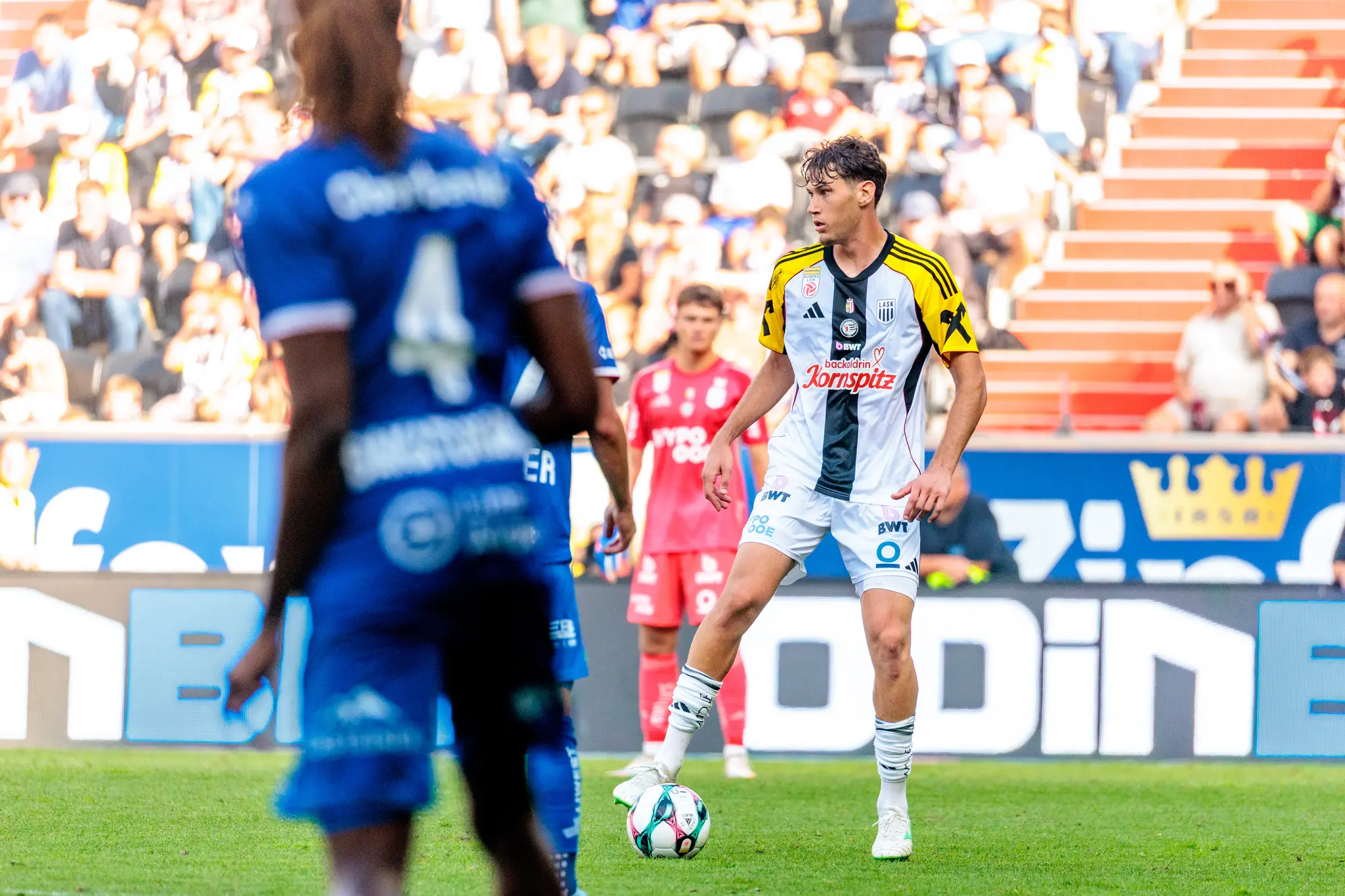 Ein Fußballspieler des LASK in weiß-schwarzem Trikot kontrolliert den Ball am Fuß, während ein Gegenspieler von Blau-Weiß Linz im unscharfen Vordergrund steht
