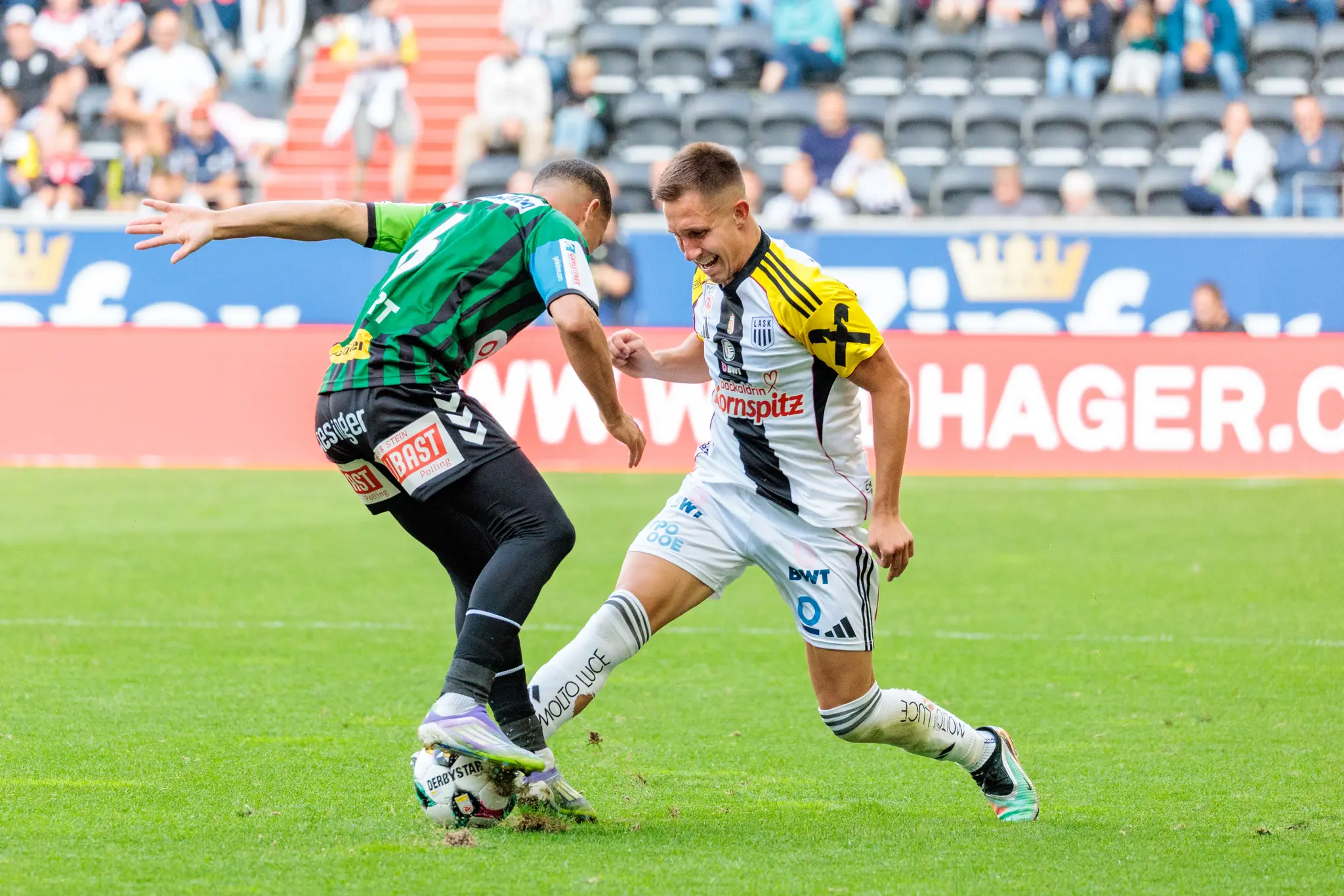 Zwei Fußballspieler, einer im weiß-schwarzen LASK-Trikot und einer im grün-schwarzen SV Ried-Trikot, liefern sich ein hartes Duell um den Ball in einem Bundesligaspiel
