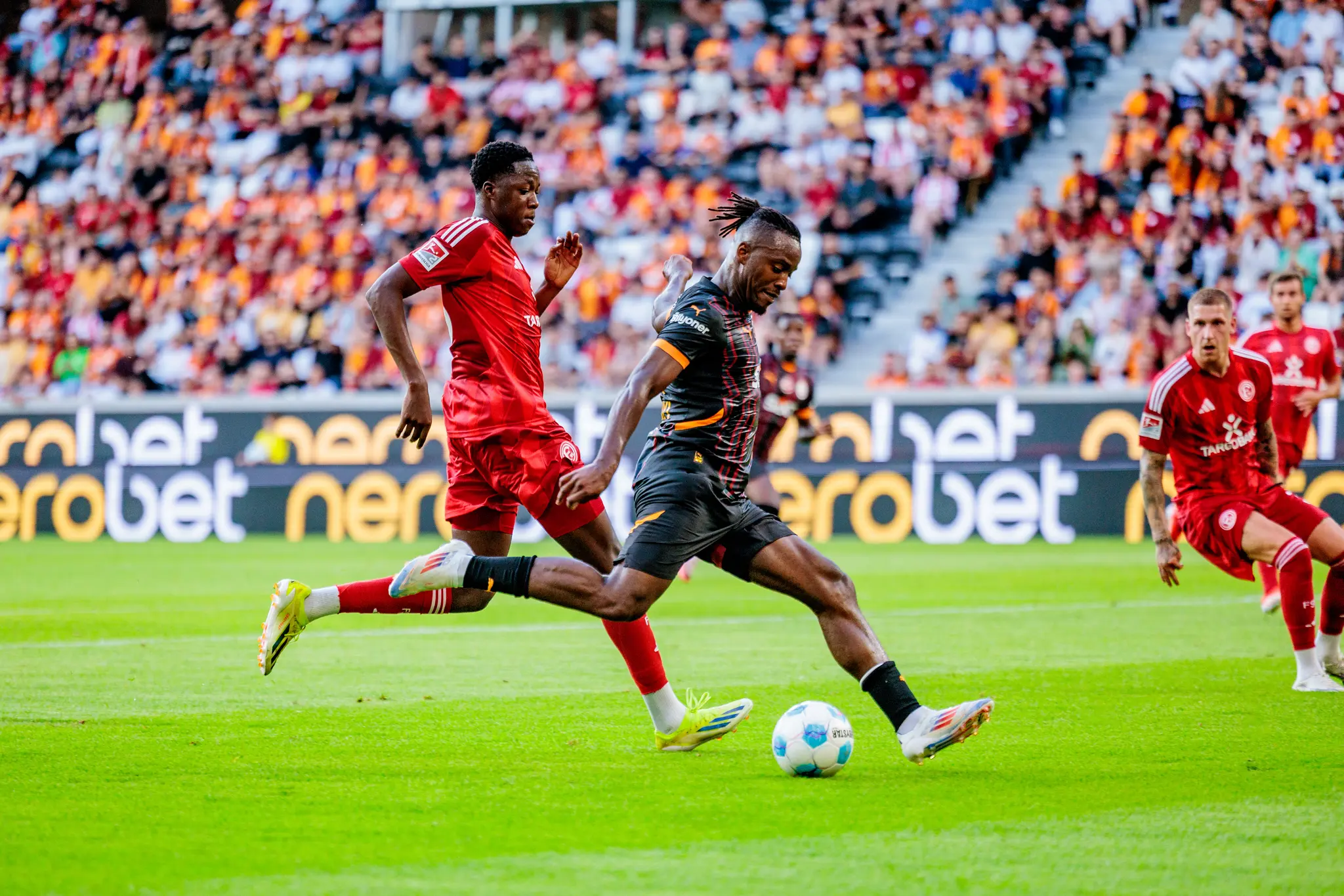 Ein Fußballspieler von Galatasaray schießt den Ball, während ein Gegner in einem roten Trikot versucht, ihn zu verteidigen 