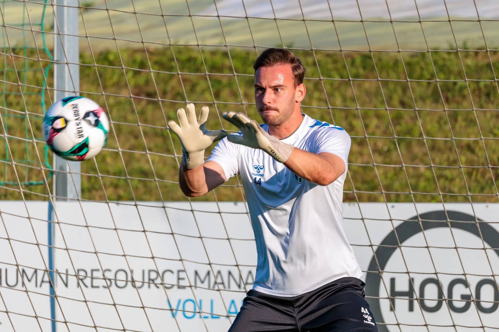 Der Torwart der SPG HOGO Wels im weißen Trikot fängt konzentriert einen Fußball beim Aufwärmen vor dem Tornetz