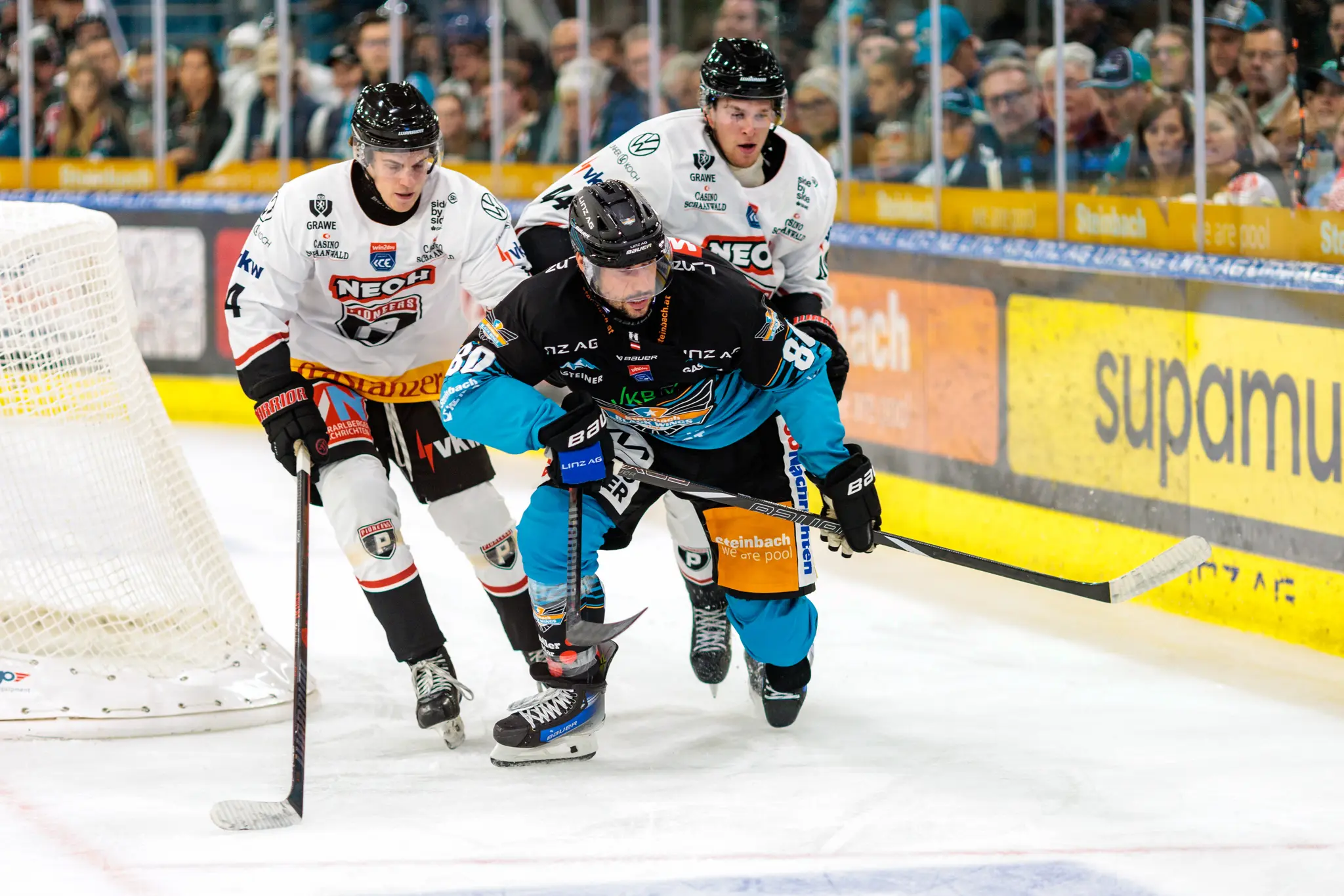 Eishockeyspieler Julian Pusnik (Nummer 80) der Steinbach Black Wings Linz skatet in seinem schwarzen Trikot mit zwei weiteren Spielern vor einem Eishockeytor auf dem Eis