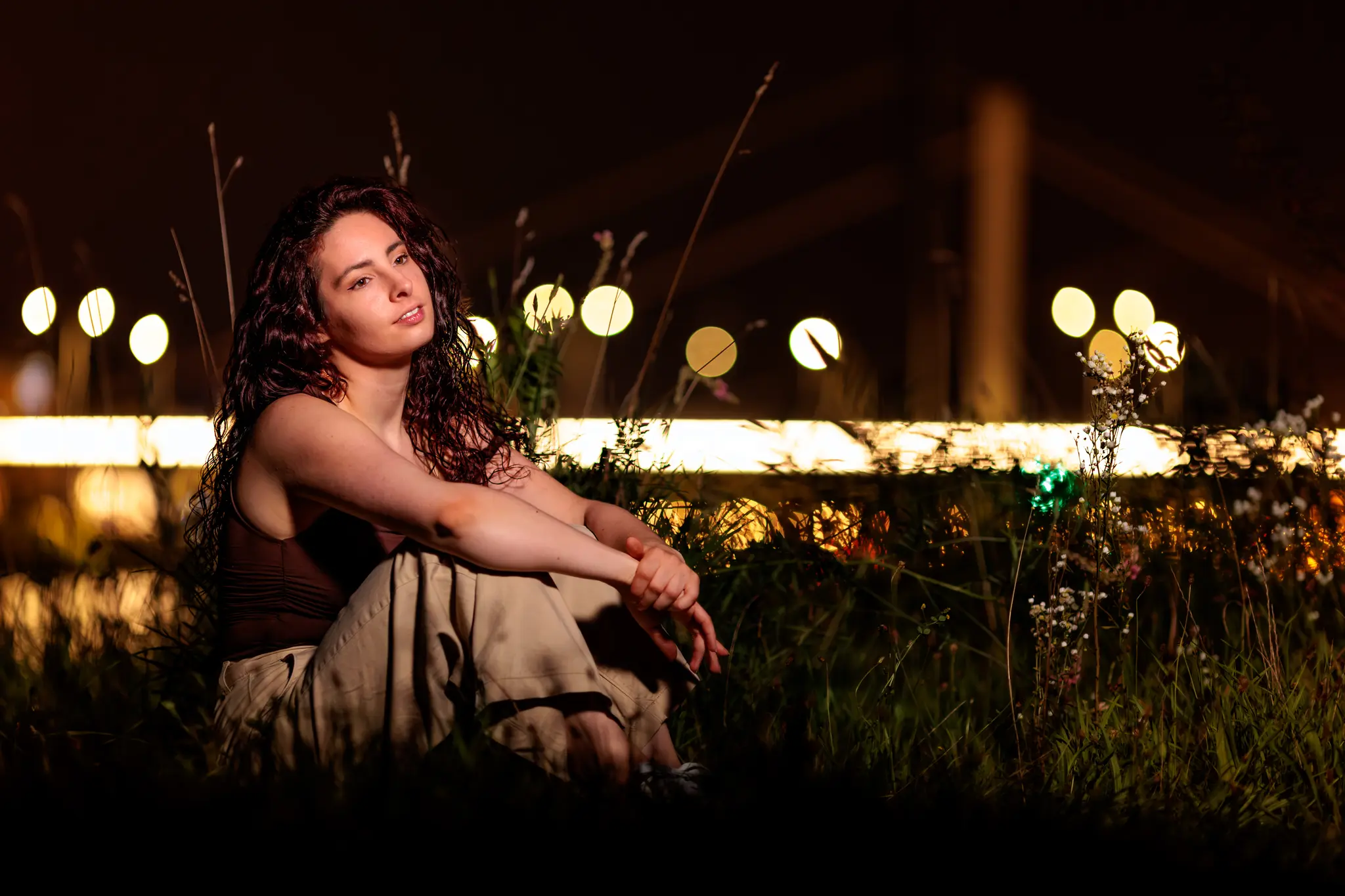Ein stimmungsvolles Nachtporträt von Nicole, die im Gras sitzt, während im Hintergrund die Lichter der Stadt Linz sanft verschwimmen