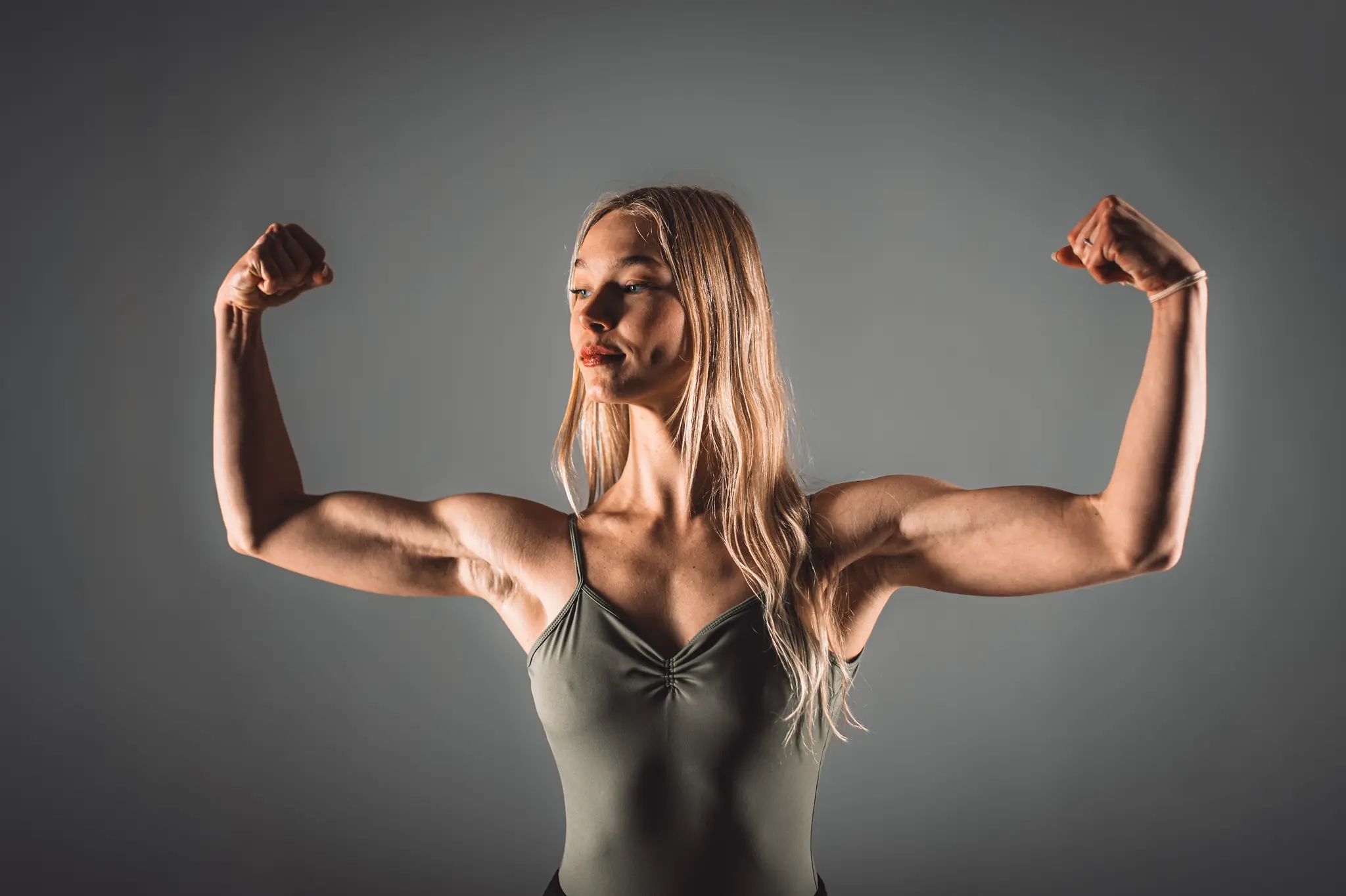 Model Kathrin präsentiert ihre beeindruckende Muskulatur und Körperbeherrschung in einer kraftvollen Bizeps-Pose vor einem schlichten grauen Hintergrund im Fotostudio
