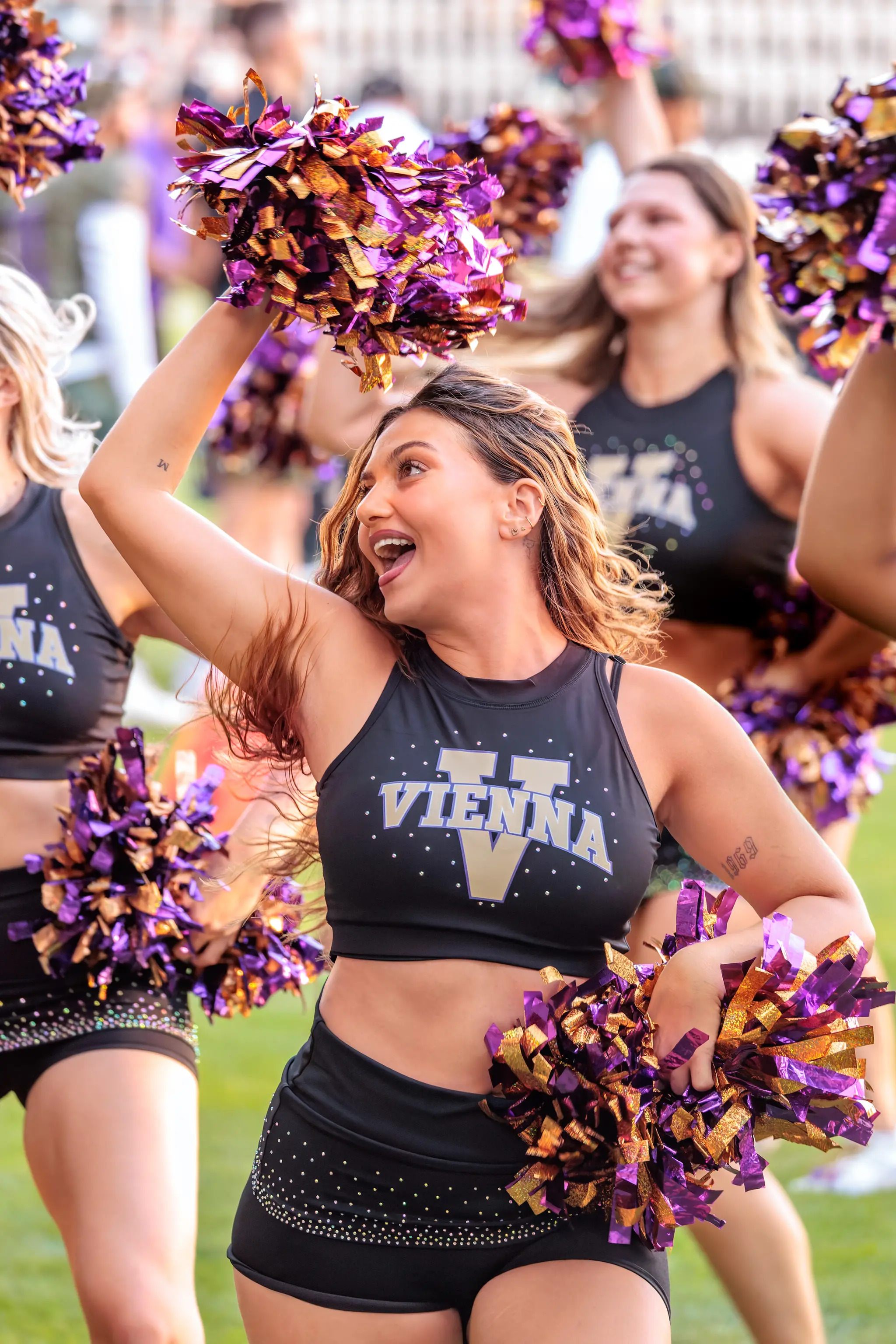 Cheerleaderin des Spirit Squad in Formation, hebt einen violett-goldenen Pompon.