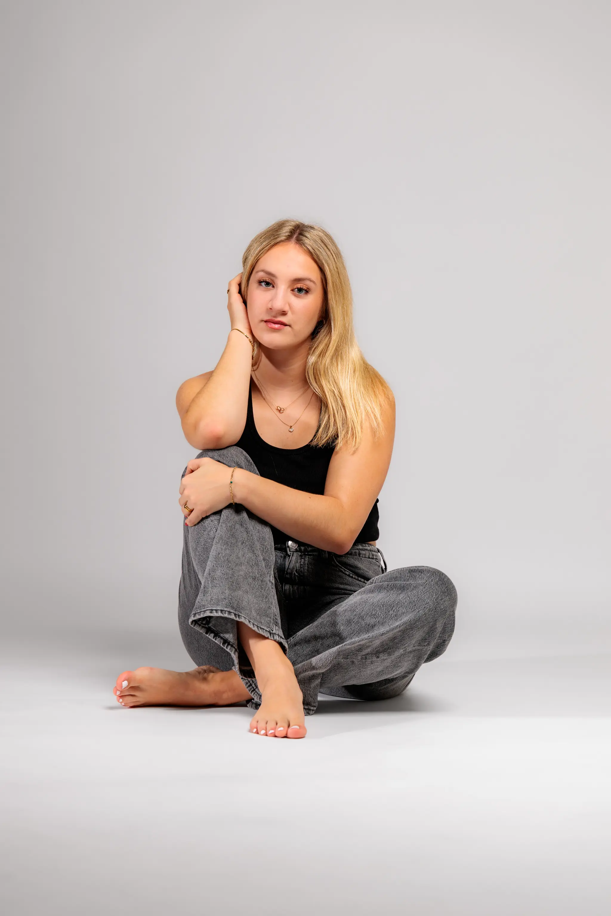 Ganzkörperaufnahme von Model Anna, die in einem lässigen Outfit mit grauer Jeans und schwarzem Top barfuß auf dem Boden des Fotostudios sitzt und in die Kamera blickt