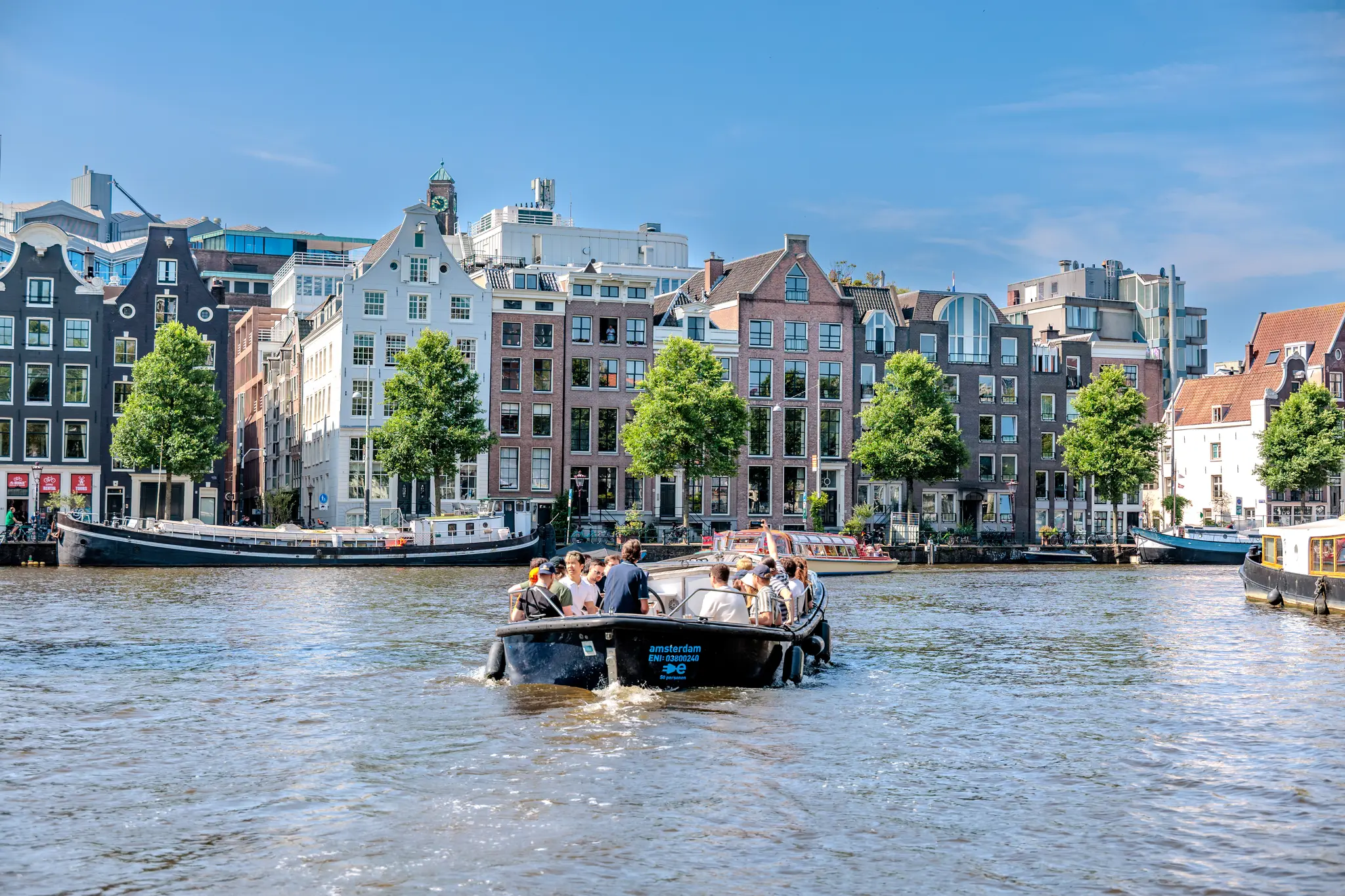 Ein Boot mit Passagieren fährt auf einem sonnigen Kanal in Amsterdam, umrahmt von den ikonischen Grachtenhäusern und Bäumen 