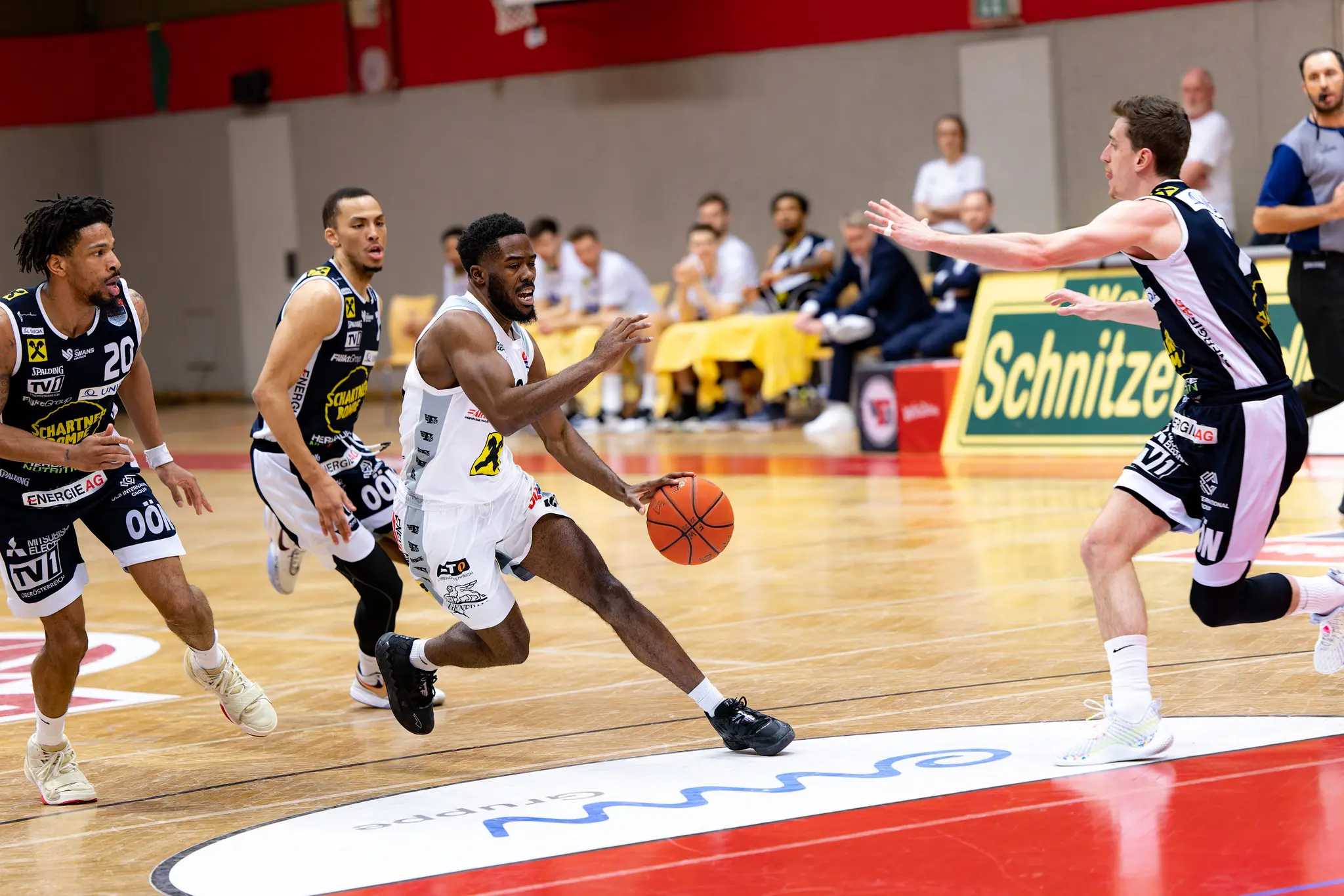 Ein Basketballspieler in einem weißen Trikot dribbelt den Ball energisch auf dem Hallenparkett, während ein Gegenspieler in dunkler Uniform ihn abwehrt