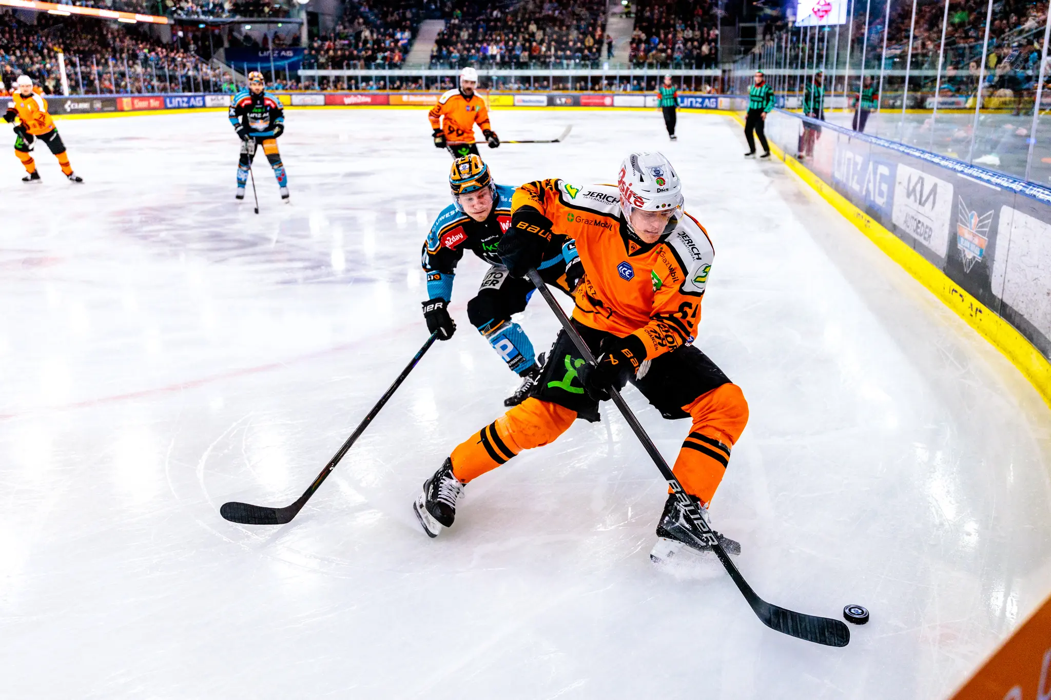 Ein Eishockeyspieler in orangefarbenem Trikot führt den Puck auf dem Eis, während ein Gegenspieler versucht, ihn zu verfolgen 