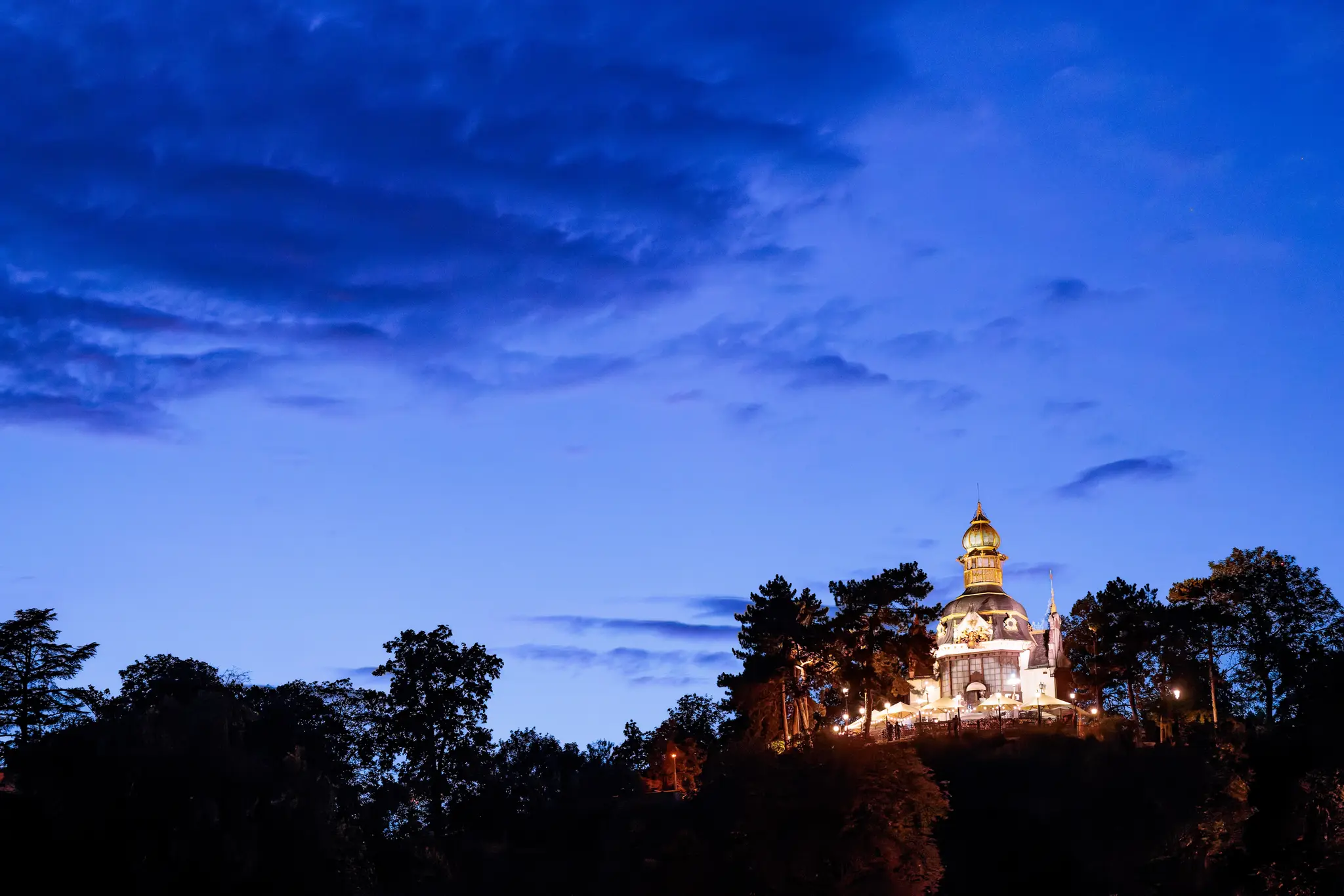 Der beleuchtete Hanavsky-Pavillon mit goldener Kuppel steht unter dramatischen Abendwolken; über den Baumkronen der Letná vertieft sich die blaue Stunde.