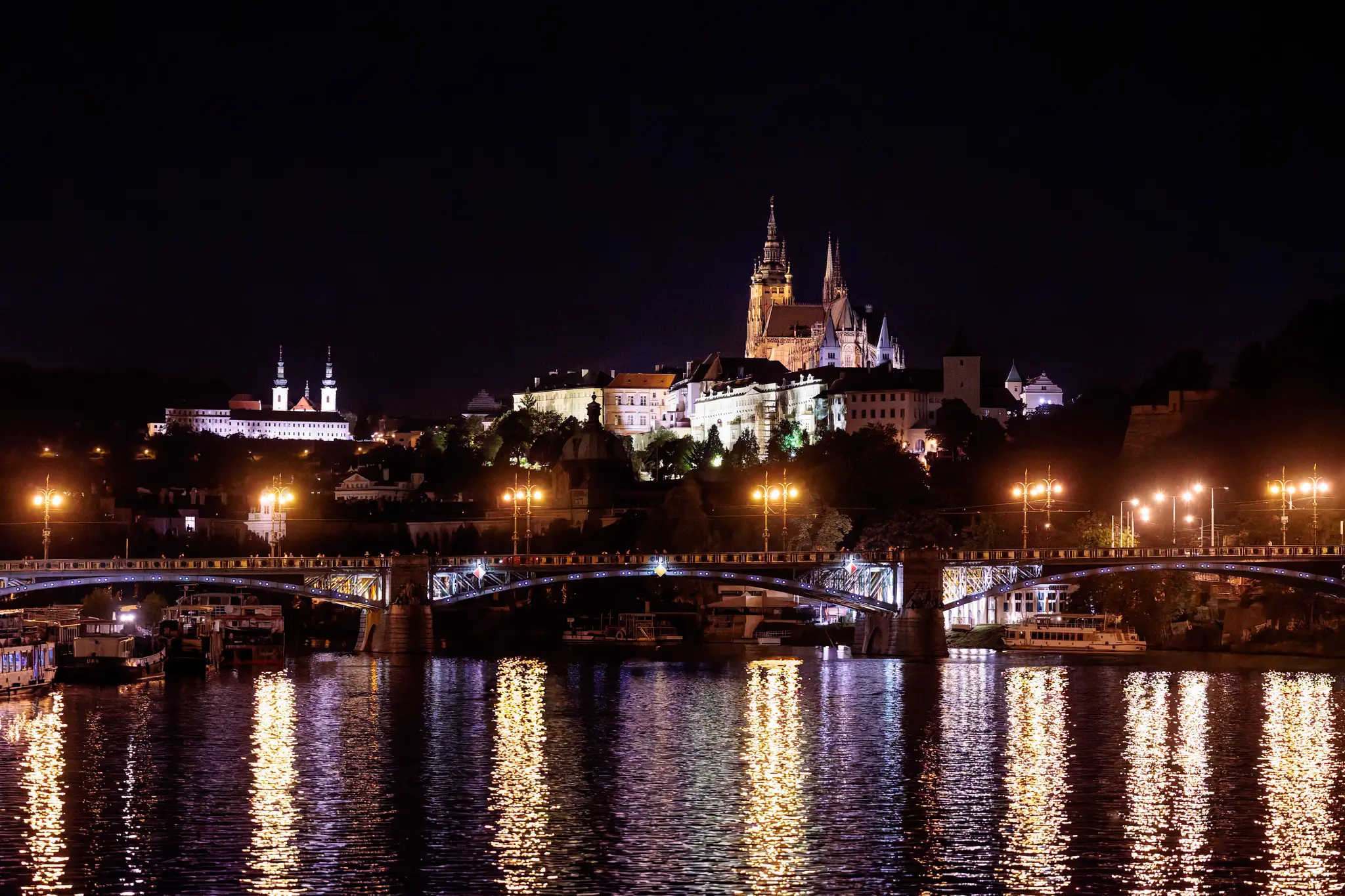 Die Prager Burg mit dem beleuchteten Veitsdom steht bei Nacht über der Moldau; Ausflugsboote und die Brücke mit Laternen spiegeln sich im Fluss.