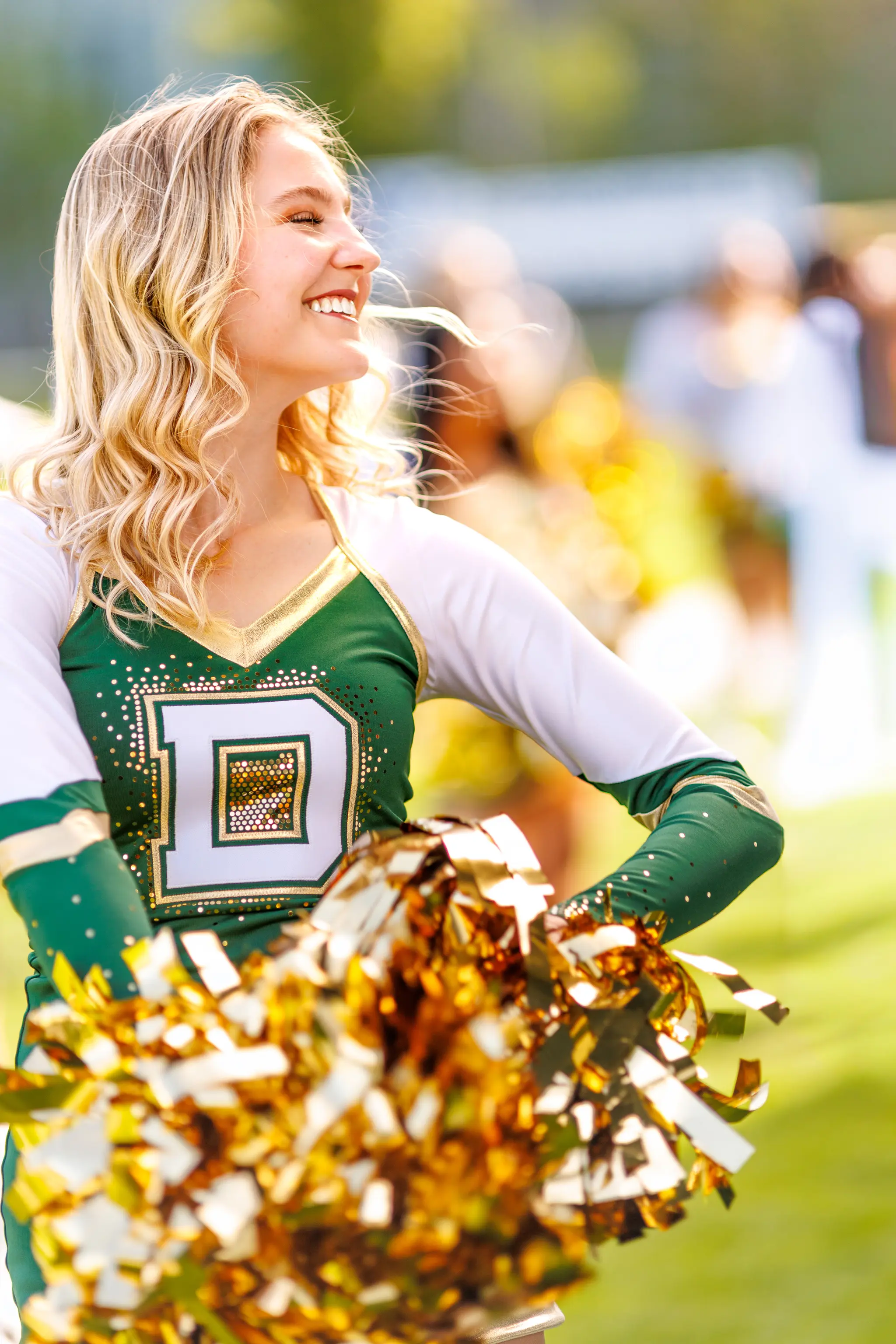 Eine lächelnde blonde Cheerleaderin in grüner und weißer Uniform hält goldene Pompons und blickt zur Seite