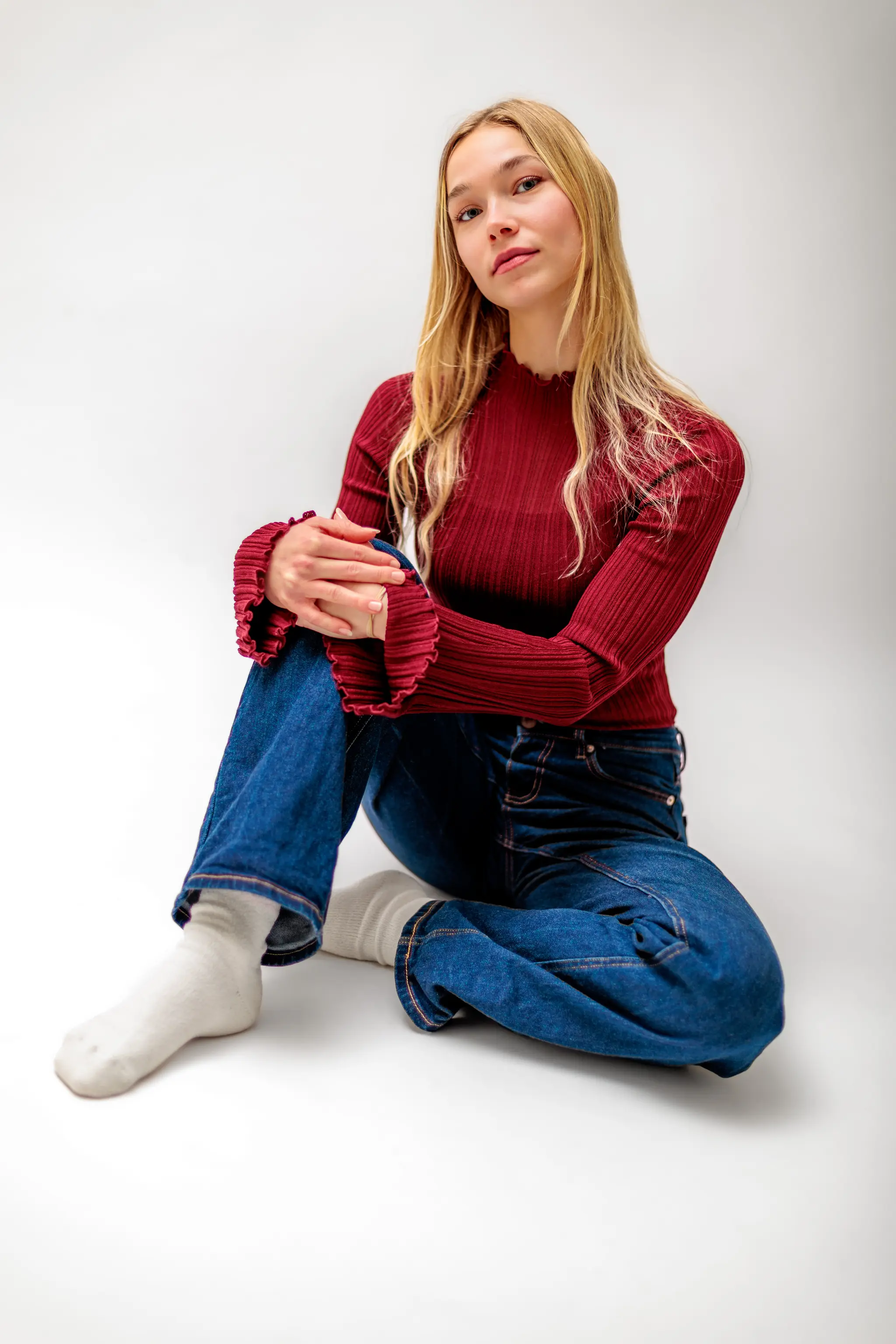 Kathrin posiert entspannt in einem bordeauxroten Strickpullover und Bluejeans für ein stilvolles Lifestyle-Portrait in einer hellen Studioumgebung