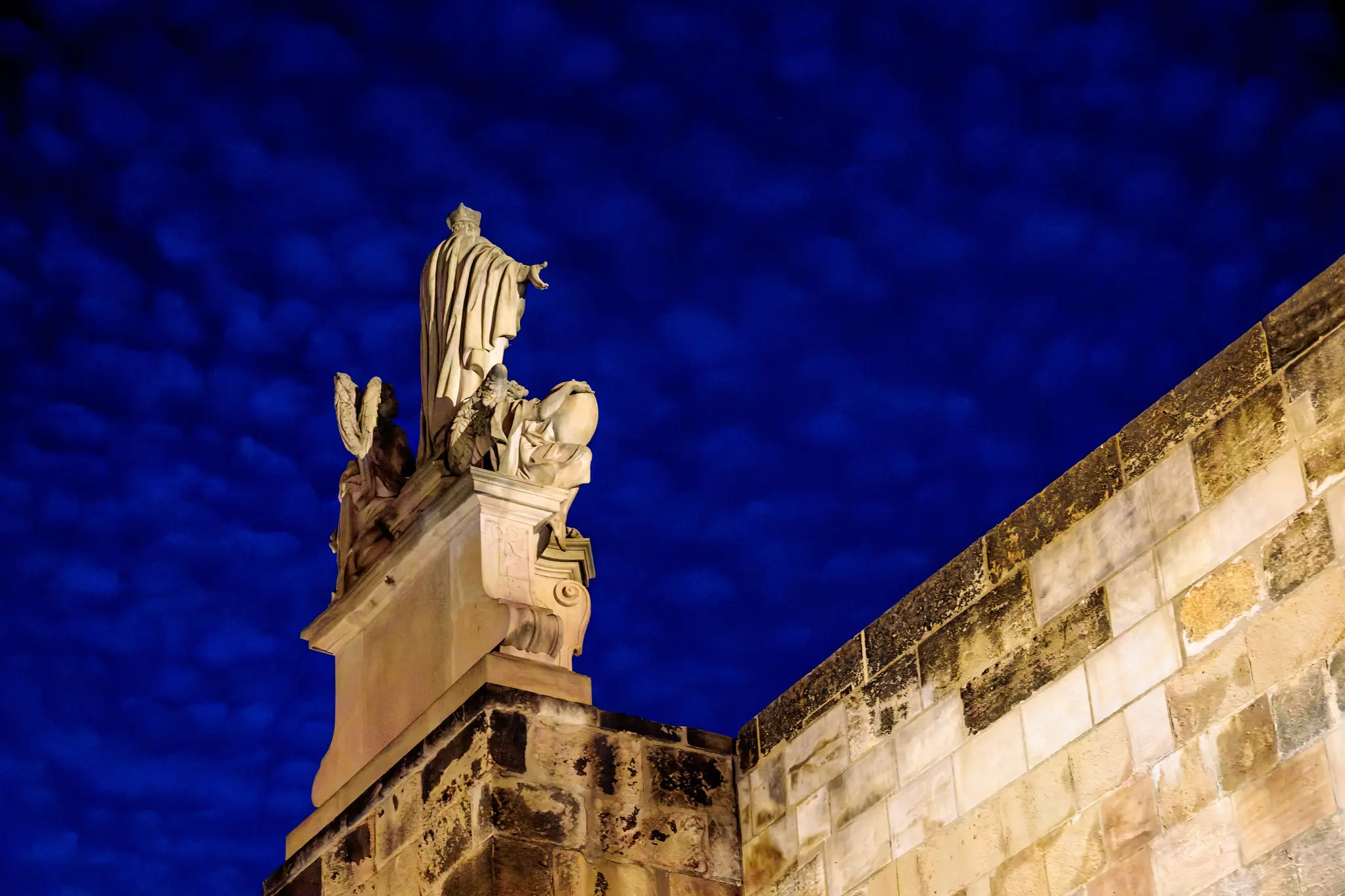 Eine Barockstatue mit Engeln thront auf dem Brückenpfeiler der Karlsbrücke; dahinter wölbt sich der tiefblaue Nachthimmel mit feinen Wolken.