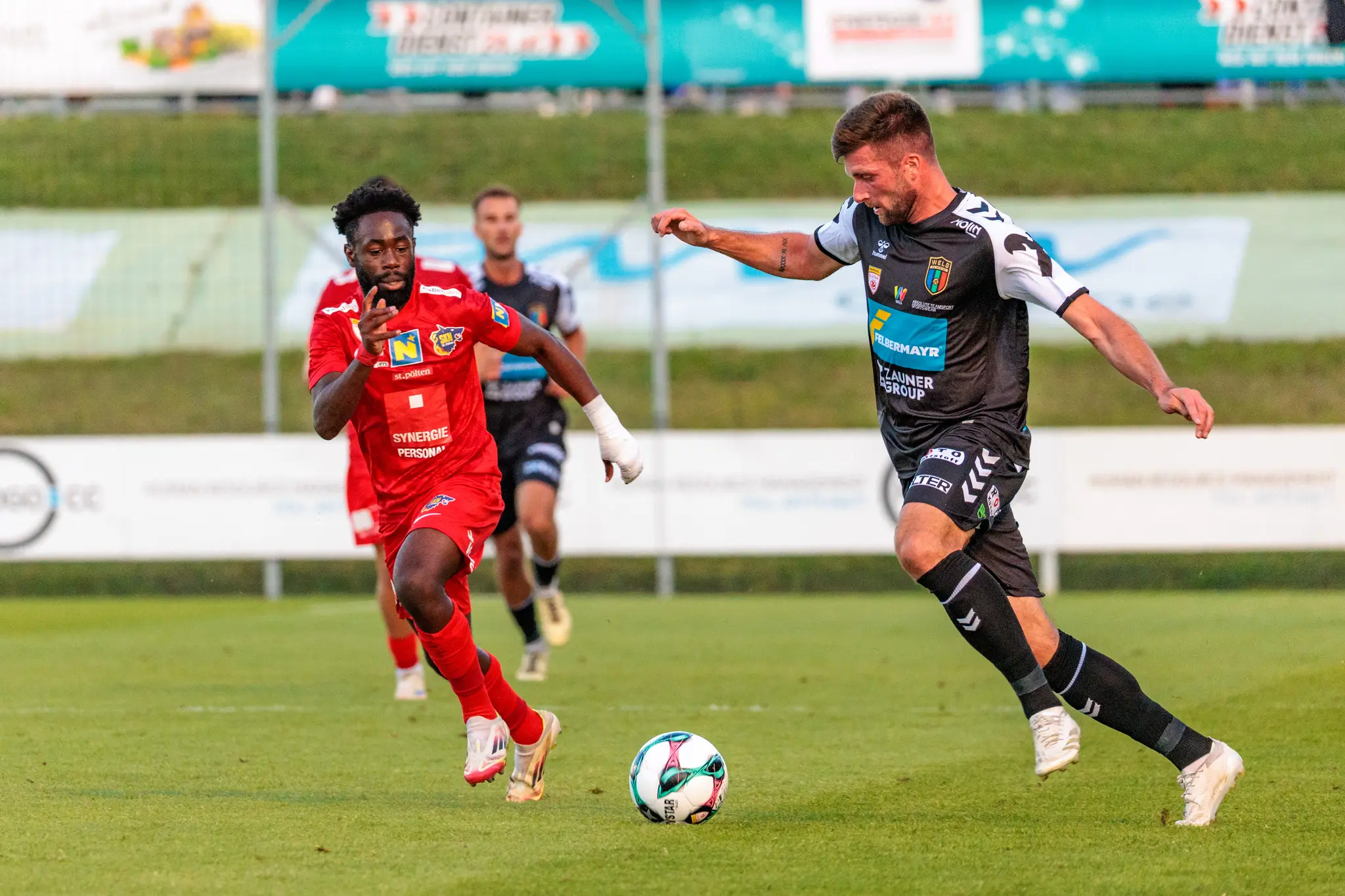 Zwei Fußballer, ein Spieler des SKN St. Pölten in rotem Trikot und ein Spieler der SPG HOGO Wels in schwarz-weißem Trikot, streiten im ÖFB-Cup-Match um den Ball