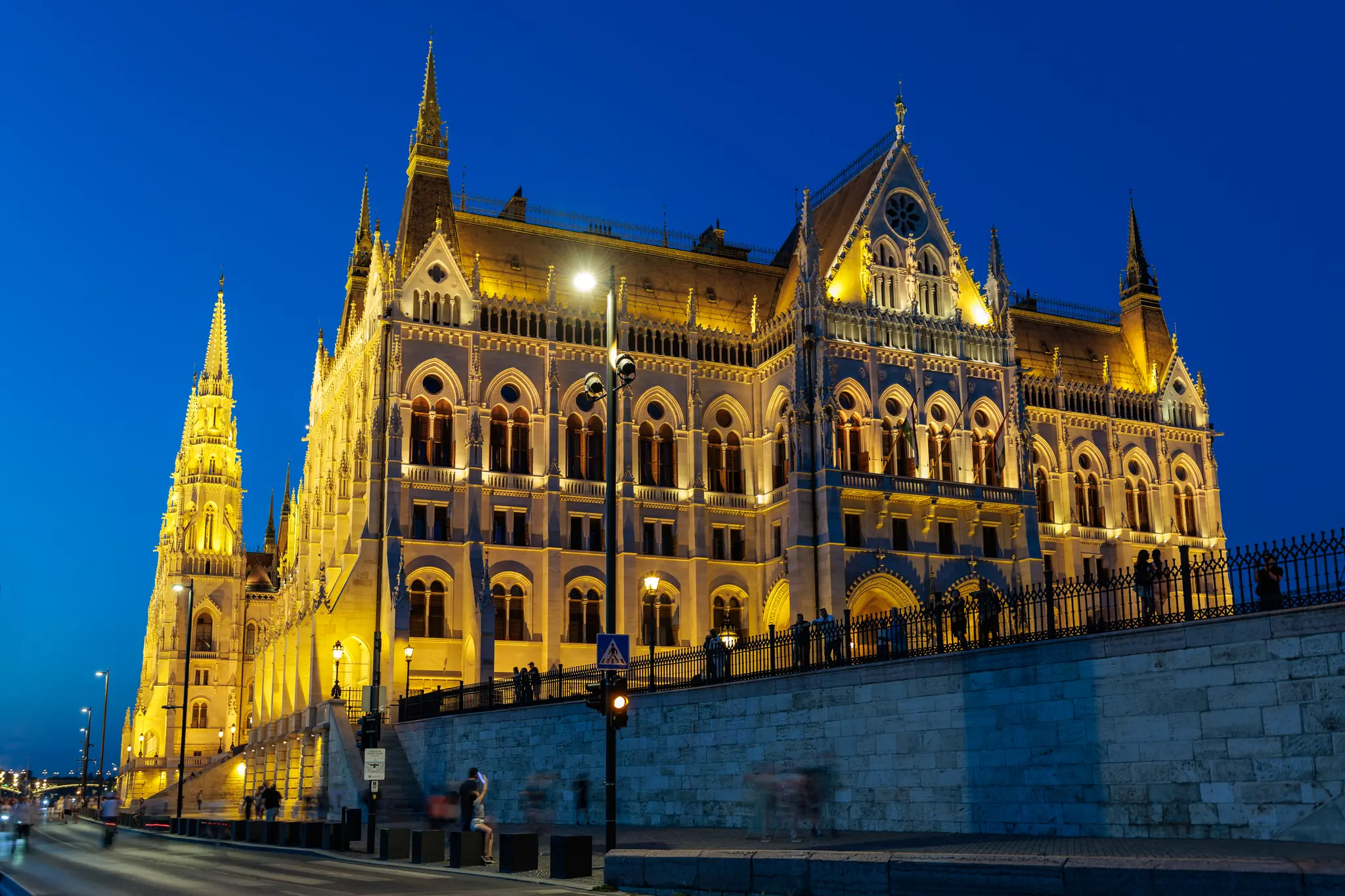 Das beleuchtete Ungarische Parlament (Országház) in Budapest bei Einbruch der Dunkelheit, mit seiner neugotischen Architektur am Donauufer und verschwommenen Menschen. 