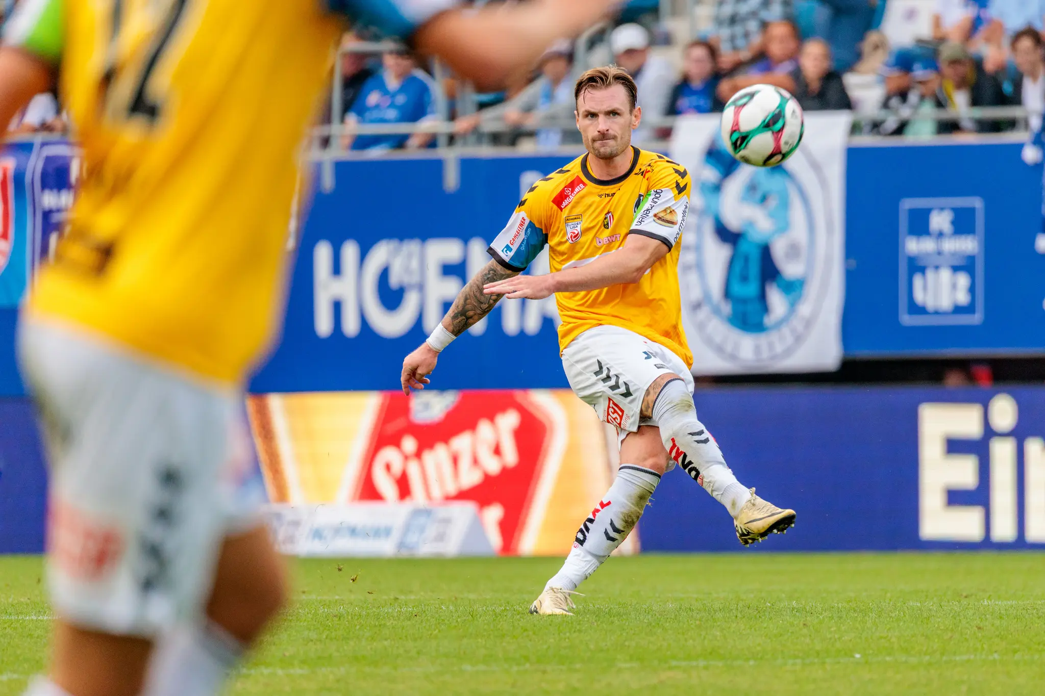 Ein SV Ried Fußballprofi im gelben Heimtrikot trifft konzentriert den Ball auf dem Feld während eines spannenden Bundesliga-Duells