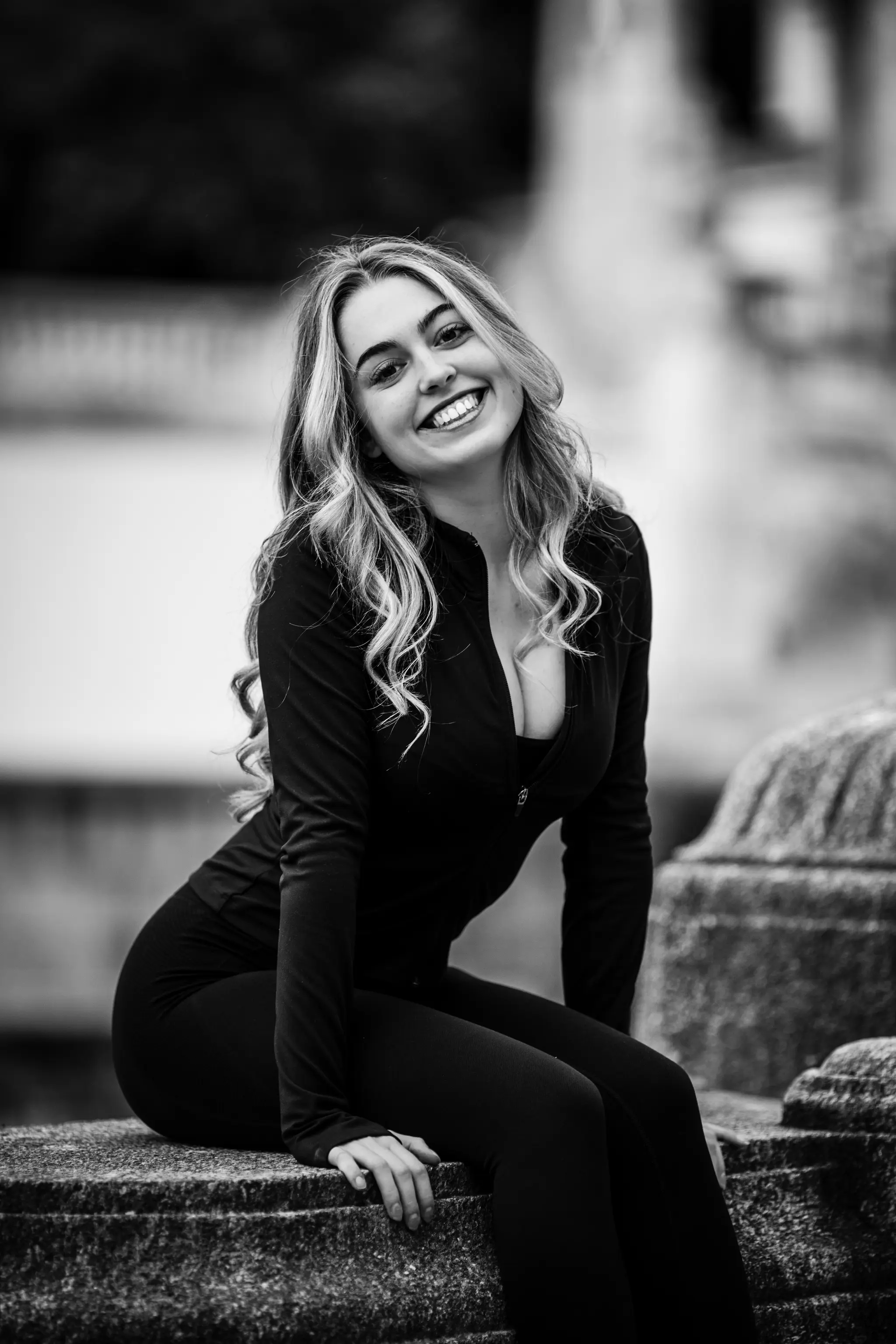 Schwarz-Weiß-Porträt von Model Valentina, die in sportlicher Kleidung auf einer Steinmauer im Stadtpark sitzt und strahlend in die Kamera lächelt