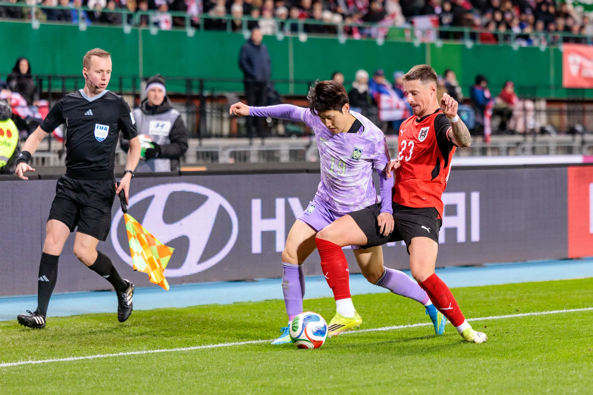 Österreichischer und südkoreanischer Spieler im intensiven Zweikampf um den Ball, überwacht von einem Schiedsrichterassistenten