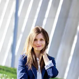 Porträt einer lächelnden Frau mit langen braunen Haaren und blauen Augen in einem dunkelblauen Blazer vor einer modernen Architekturkulisse mit markanten diagonalen Licht- und Schattenlinien