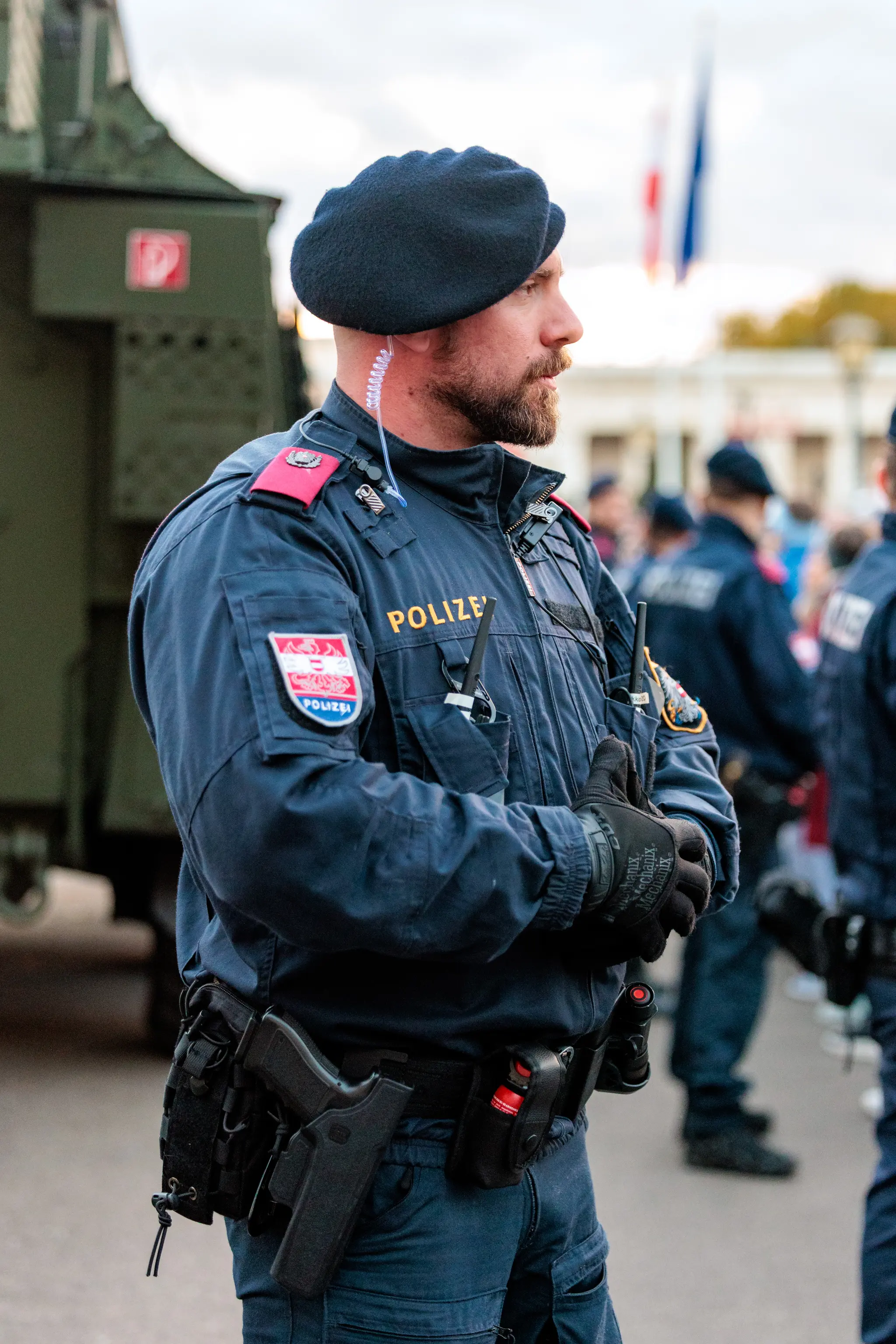 Ein Polizist mit blauem Baret und dunkelblauer Einsatzjacke steht im Profil vor einem Militärfahrzeug, die Hände vor dem Körper verschränkt.