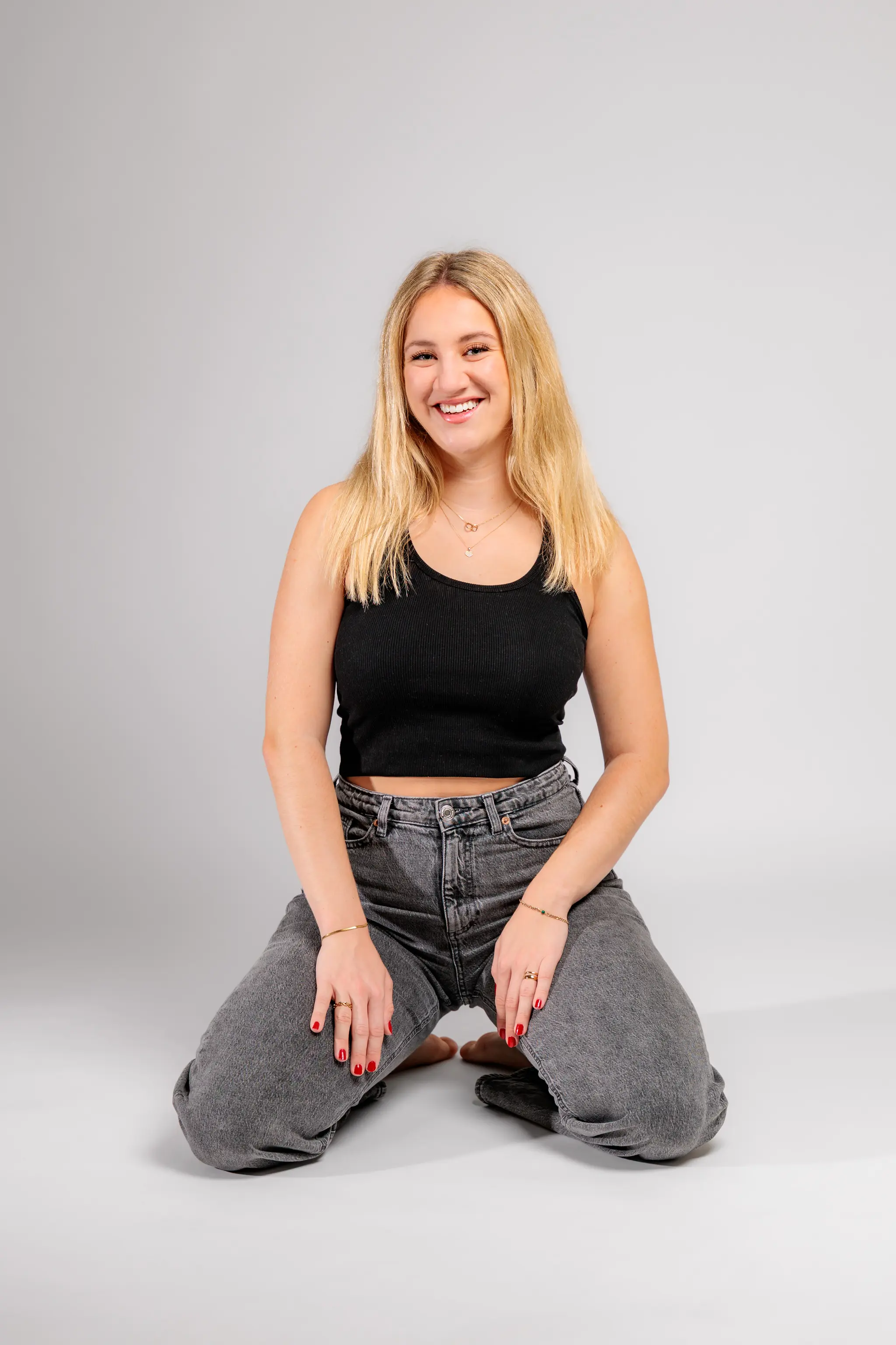 Das Model Anna kniet in einem lässigen Outfit mit Jeans und schwarzem Top auf dem weißen Boden des Fotostudios und lächelt in die Kamera