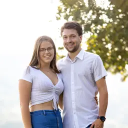Porträtfoto von einem lächelnden jungen Mann und einer jungen Frau, die im Freien vor einem sonnenbeschienenen, unscharfen Grün stehen lens: 56mm F1.4 DC DN | Contemporary 018