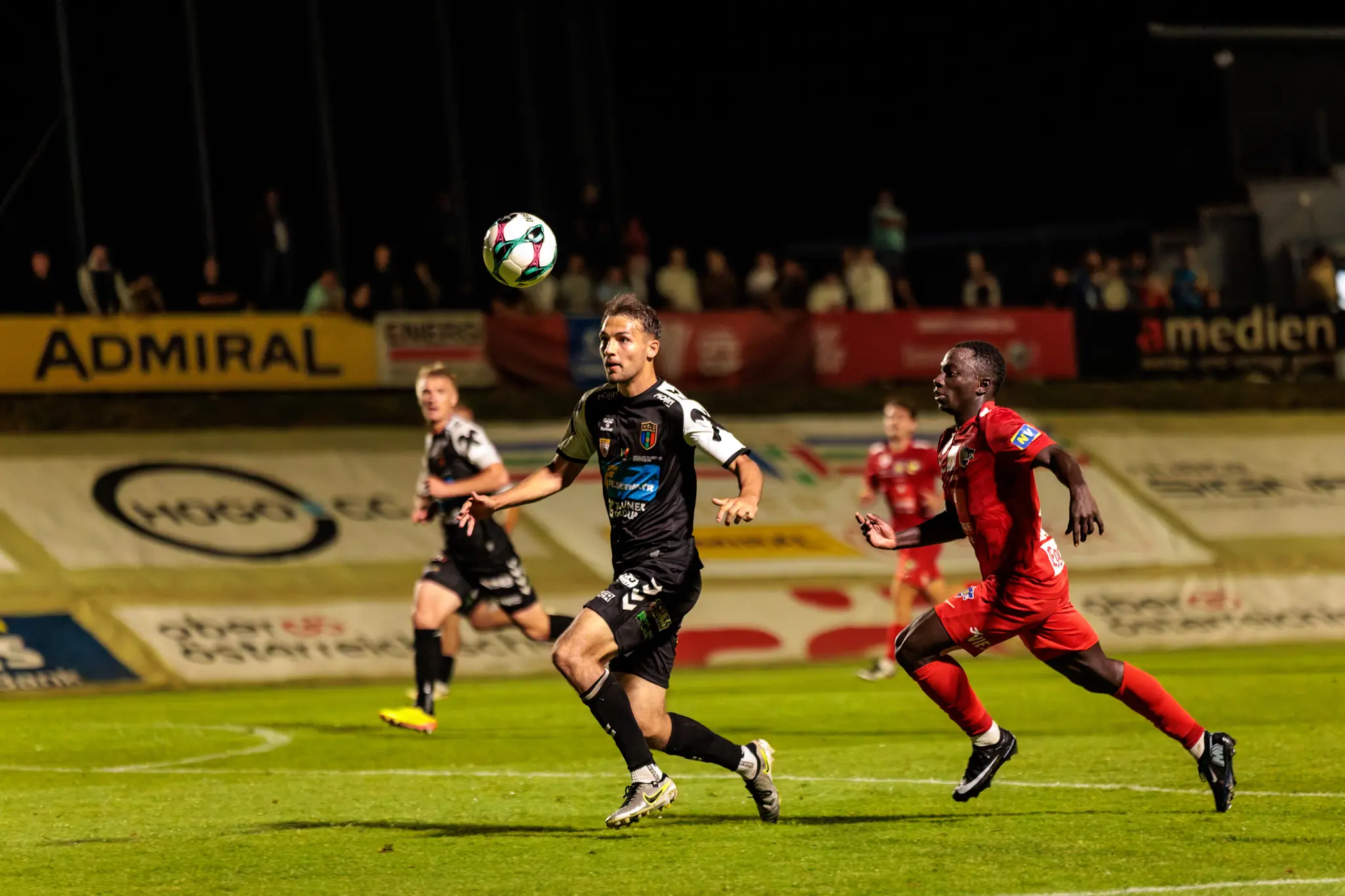 Ein Spieler in schwarzem Trikot und ein Spieler in rotem Trikot liefern sich ein intensives Laufduell um den Ball bei einem Flutlicht-Pokalspiel