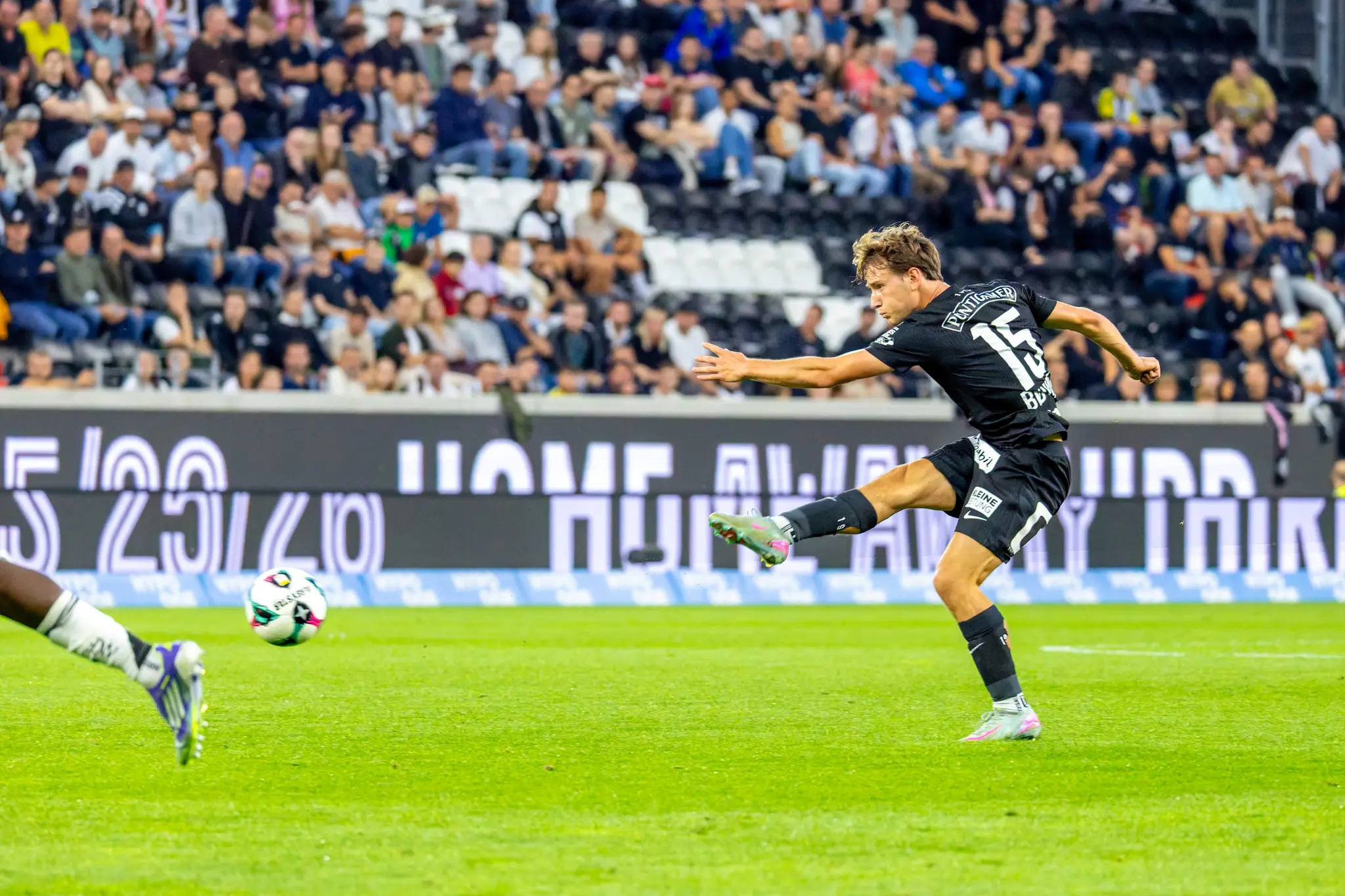 Ein Fußballspieler in schwarzem Trikot mit der Nummer 15 ist in voller Schussbewegung vor unscharfen Zuschauern