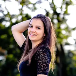 Eine lächelnde junge Frau mit langen braunen Haaren und schwarzem Spitzenoberteil posiert im Freien vor einem unscharfen grünen Hintergrundlens: 56mm F1.4 DC DN | Contemporary 018