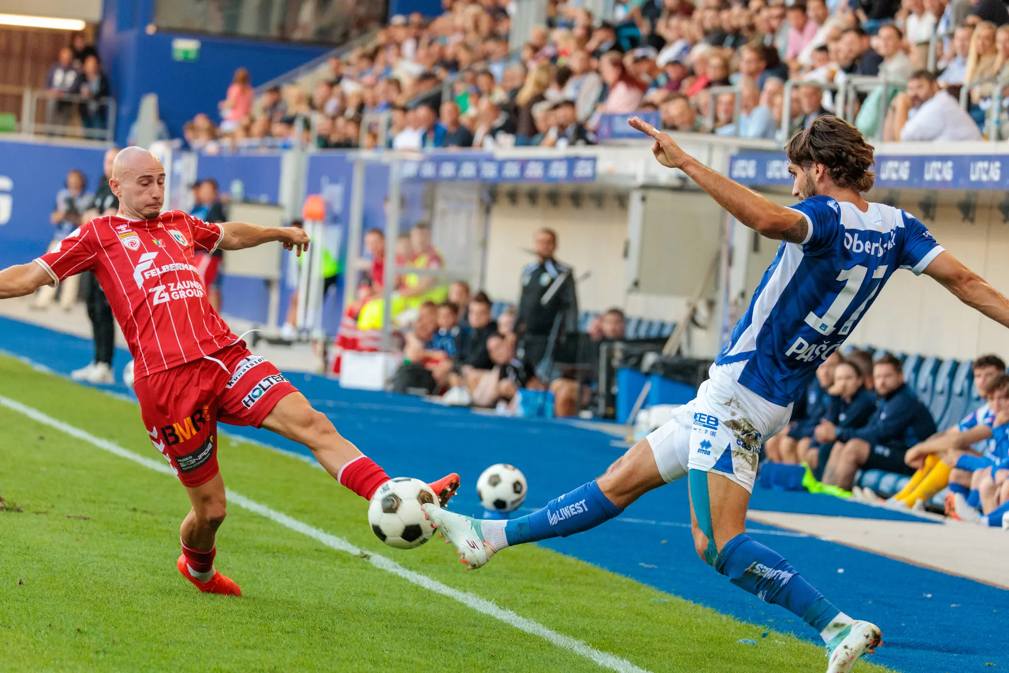 Alem Pasic vom FC Blau-Weiß Linz geht an der Seitenlinie in den Zweikampf mit einem Welser, der den Ball wegspitzelt; im Hintergrund sind Ersatzbank und Tribüne zu sehen.