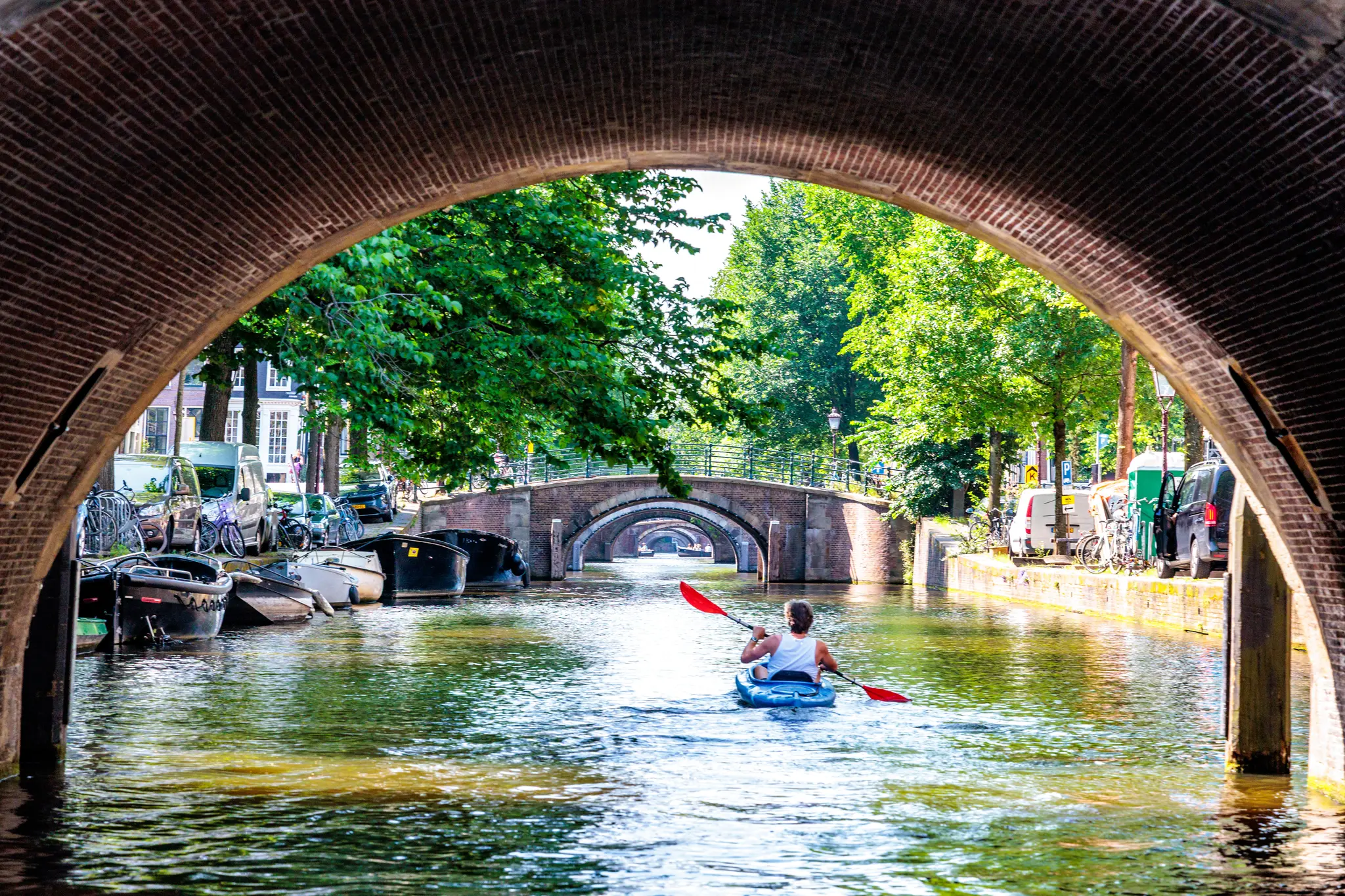 Eine Person in einem blauen Kajak paddelt unter einer historischen Backsteinbrücke auf einem sonnigen Kanal in Amsterdam, umgeben von grünen Bäumen und urbaner Architektur 